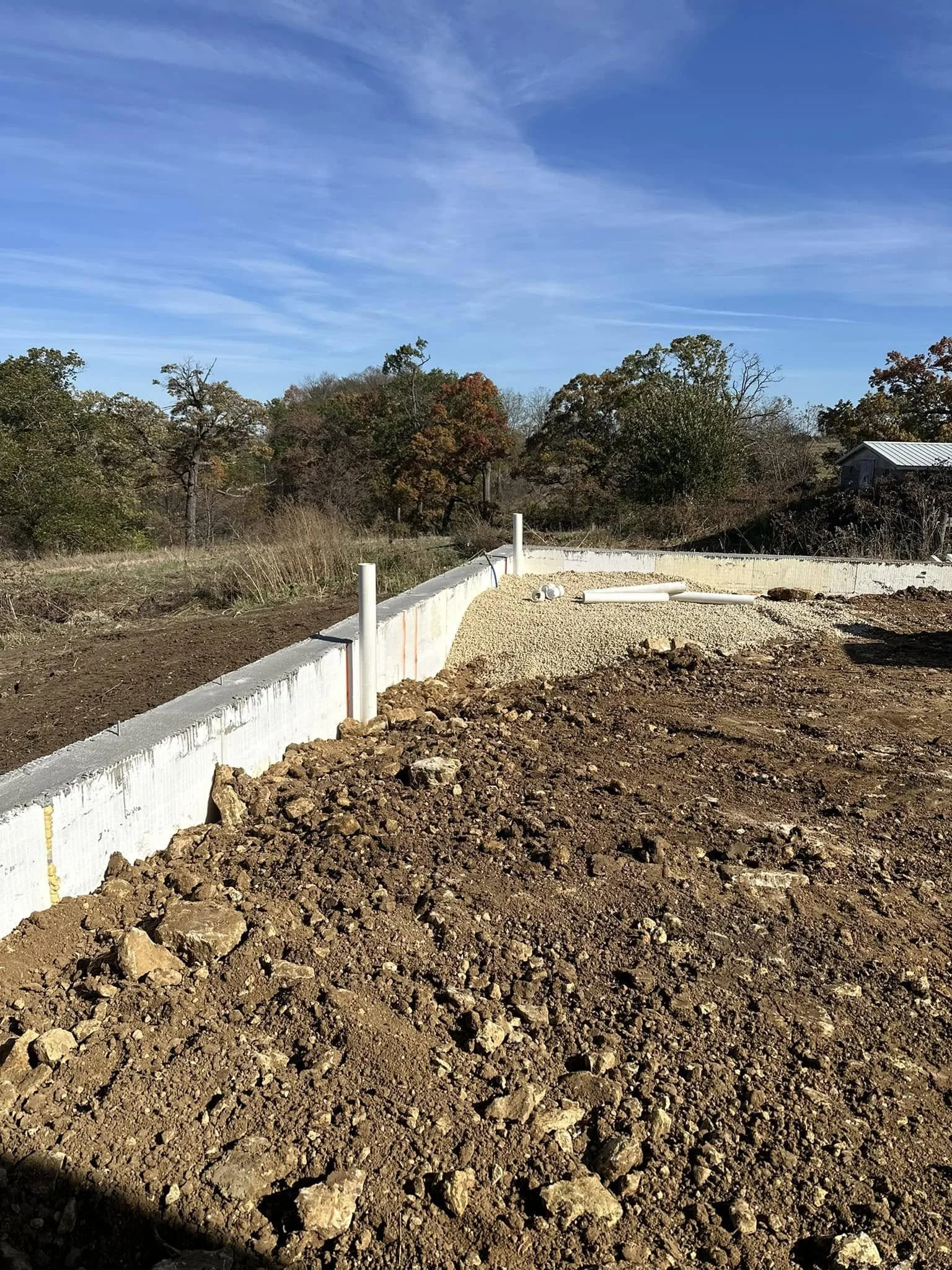 Construction site with a concrete foundation and pipes, surrounded by brown dirt, with trees and a blue sky in the background.