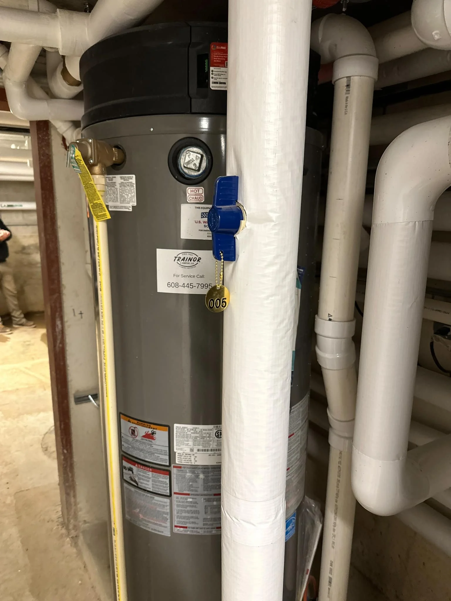 A water heater tank surrounded by white pipes in a basement or utility room.