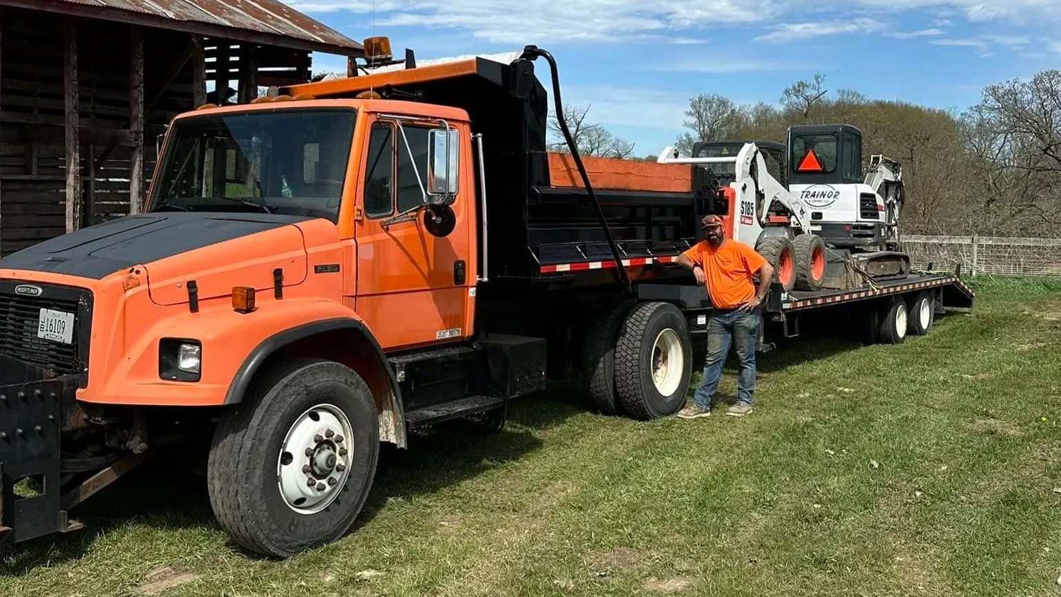 A person standing beside a flatbed tow truck with a skid-steer loader on it, outdoors on grass with a fence and trees in the background.