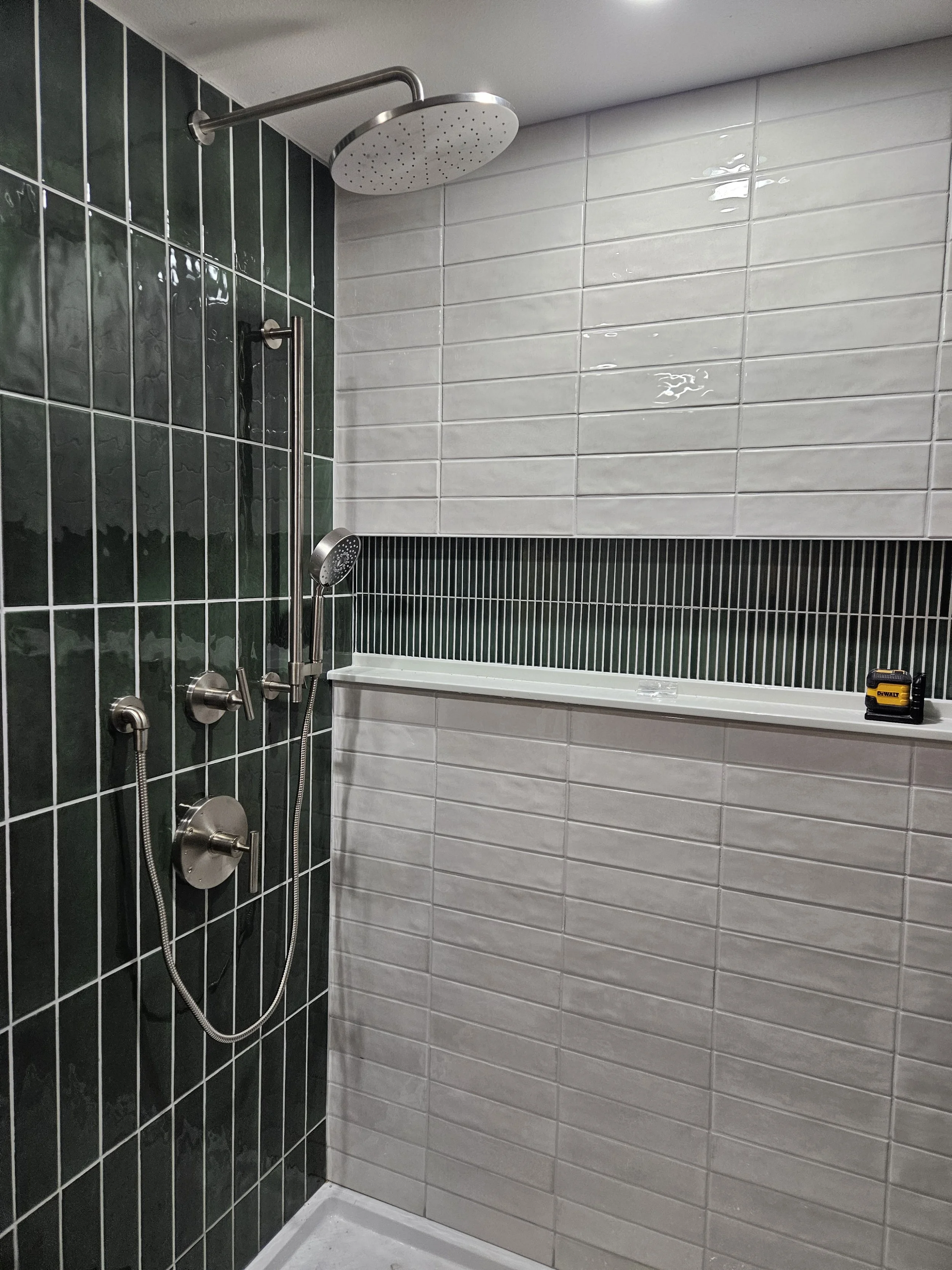 Corner of a bathroom shower with black and white tiles, a large overhead showerhead, a handheld showerhead, and a small tape measure on the ledge.