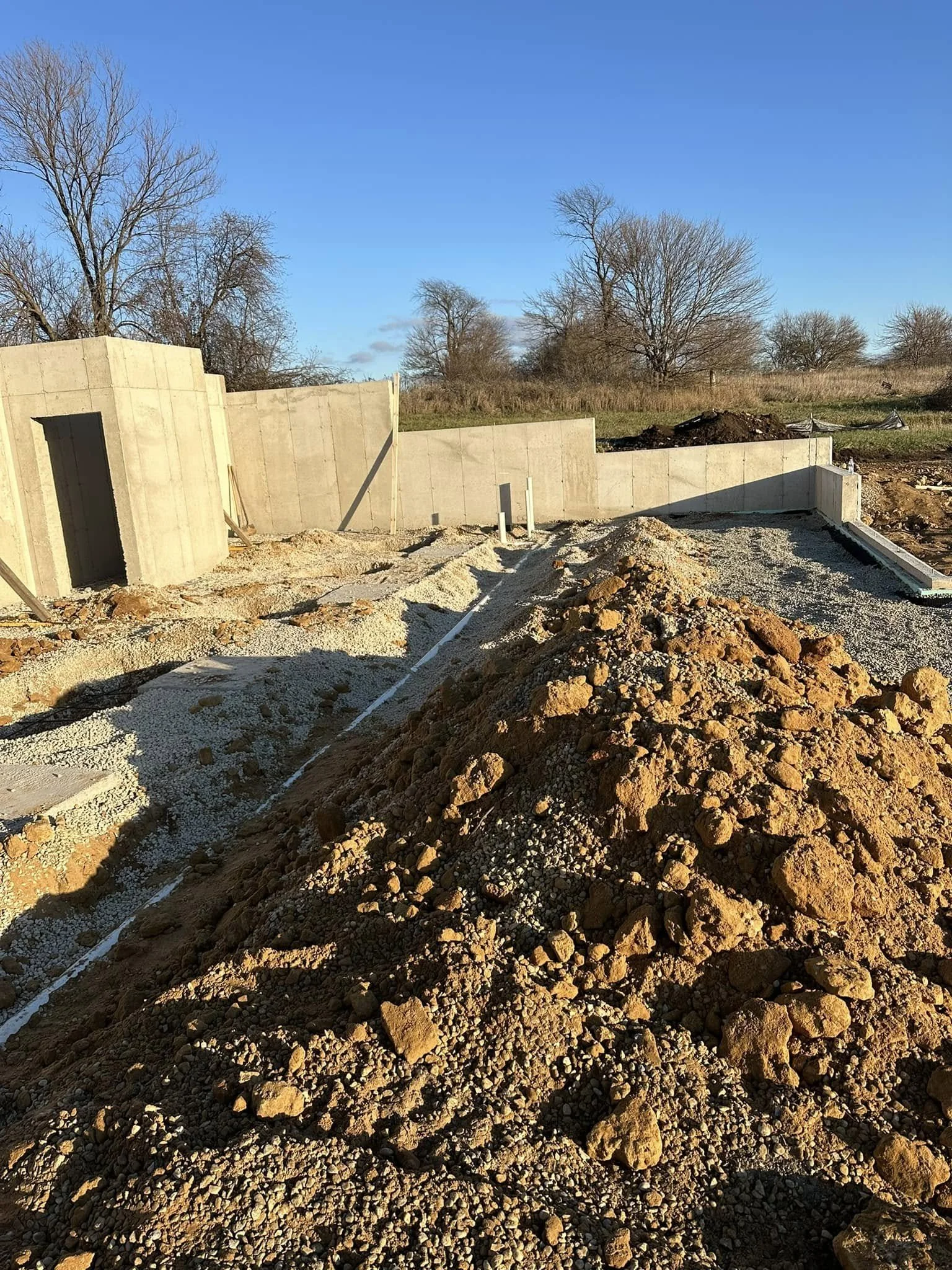 Construction site with dirt piles and unfinished concrete walls under a clear blue sky, with leafless trees in the background.
