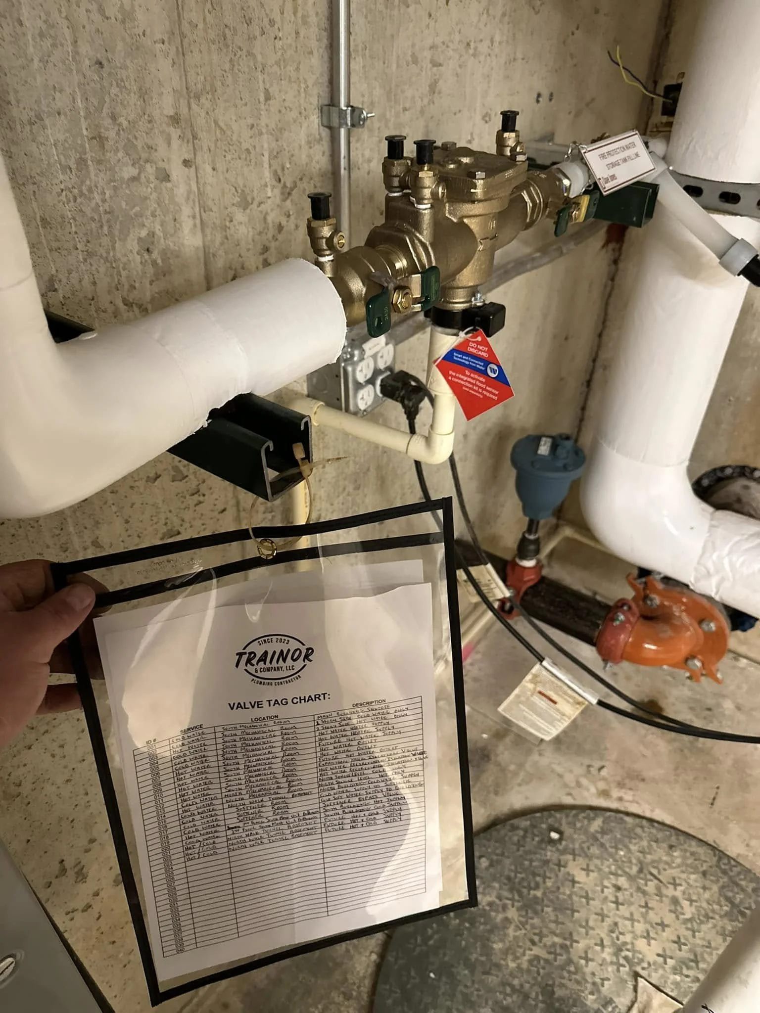 A plumbing valve tag chart held in front of a plumbing pipe system, with pipes, valves, and electrical wires visible in the background.