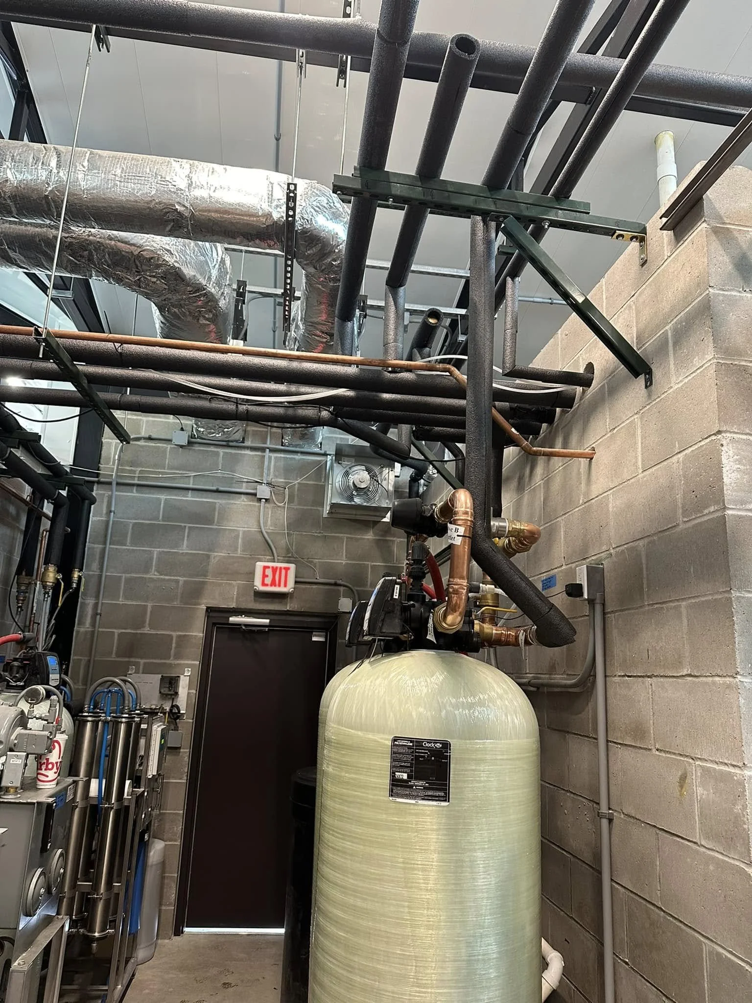 View of industrial pipes, a water tank, and mechanical equipment in a utility room with brick walls and an exit sign on the wall.