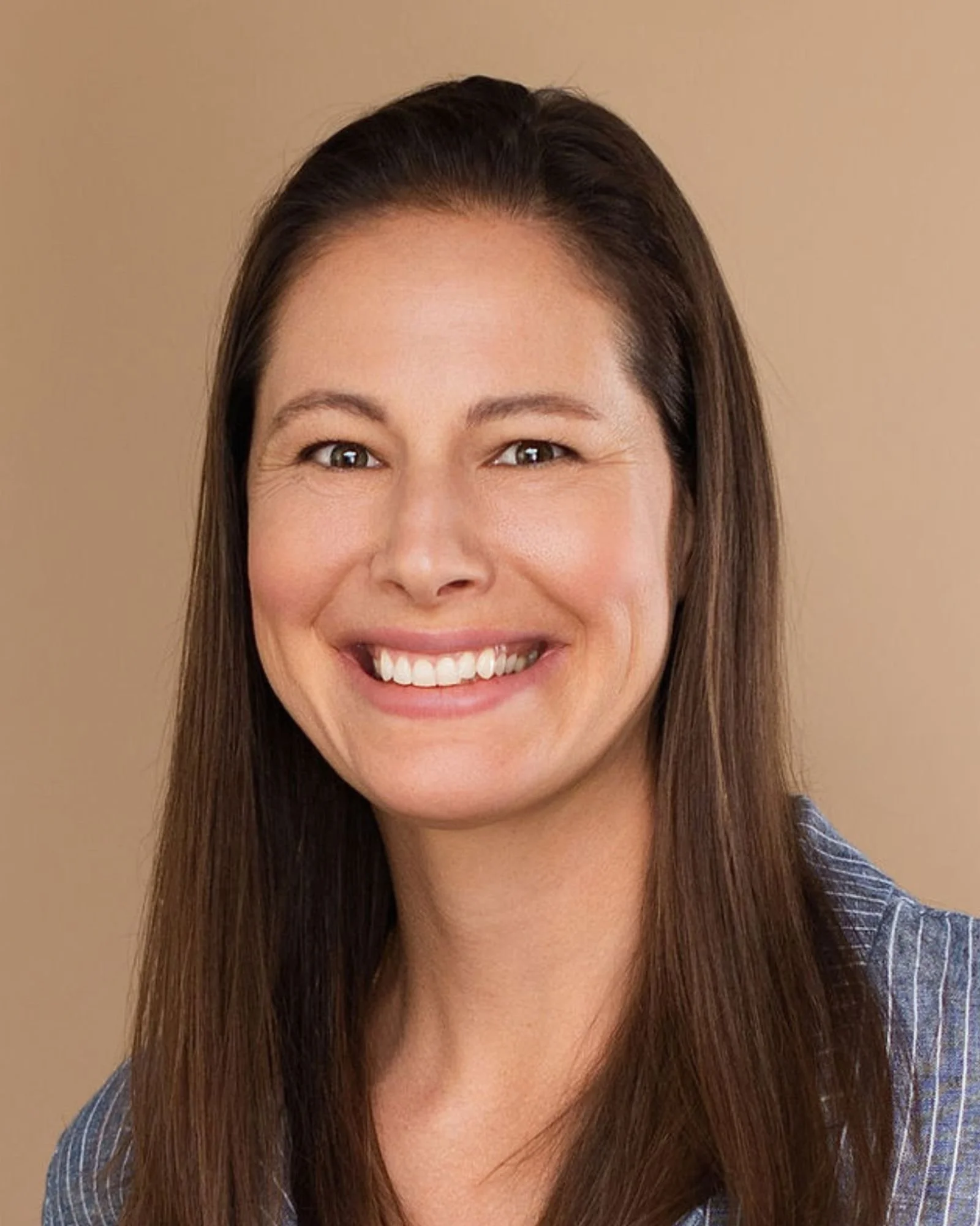 Close-up of a woman with long brown hair smiling and showing her teeth, wearing a striped shirt, against a plain beige background.