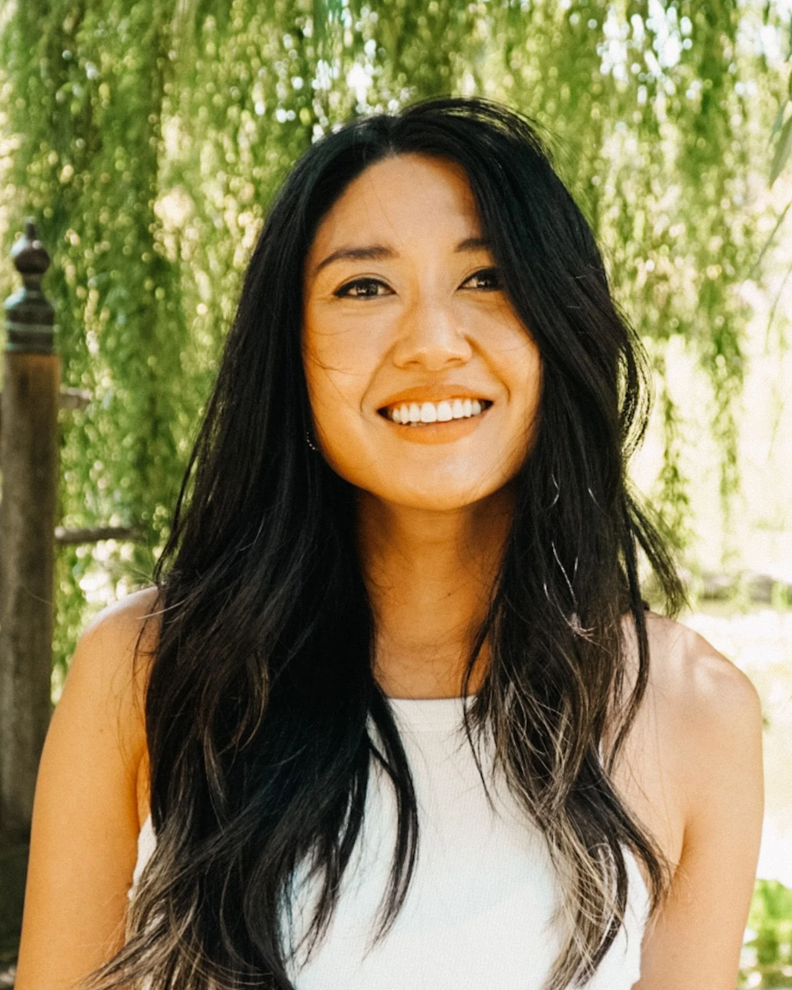 A woman with long black and gray hair smiling outdoors with green trees in the background.