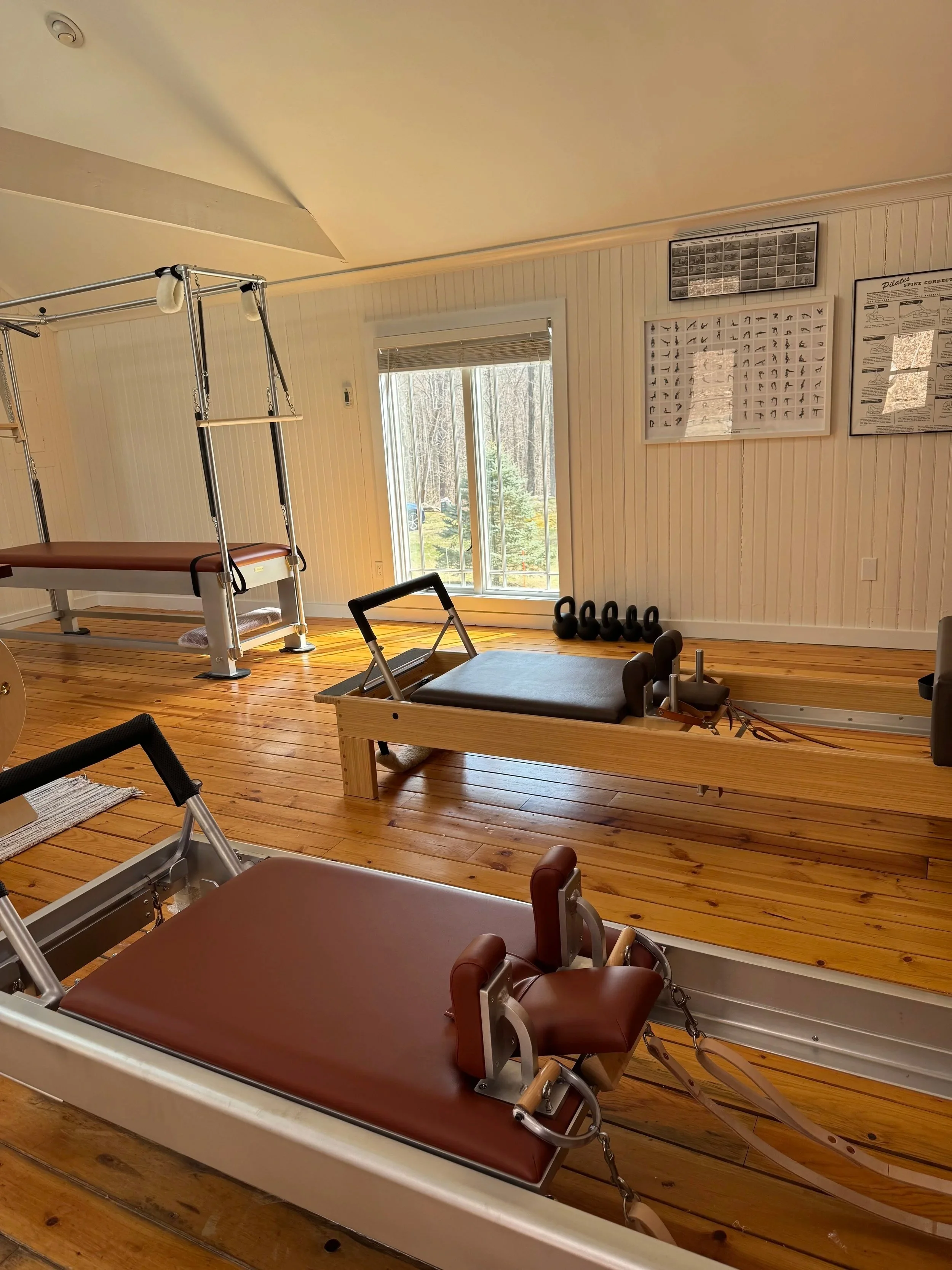 Pilates cottage gym with wooden floors, featuring  a Cadillac, two Pilates reformers, kettlebells, and posters on the wall near a large window.