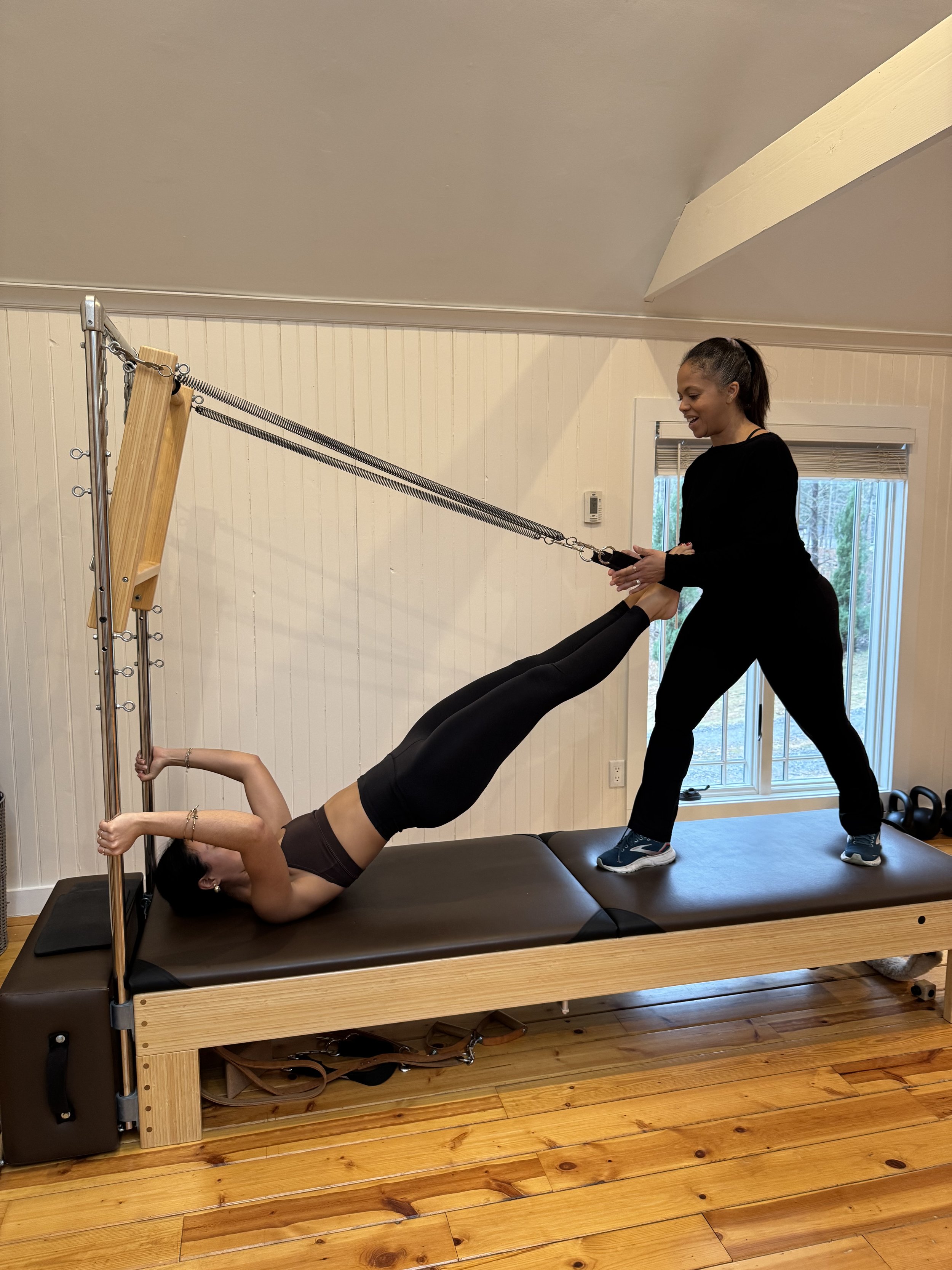 A woman lying on a Tower reformer Pilates machine, stretching with her arm on the poles, legs extended while holding onto the machine's spring and foot straps, and another woman assisting her.