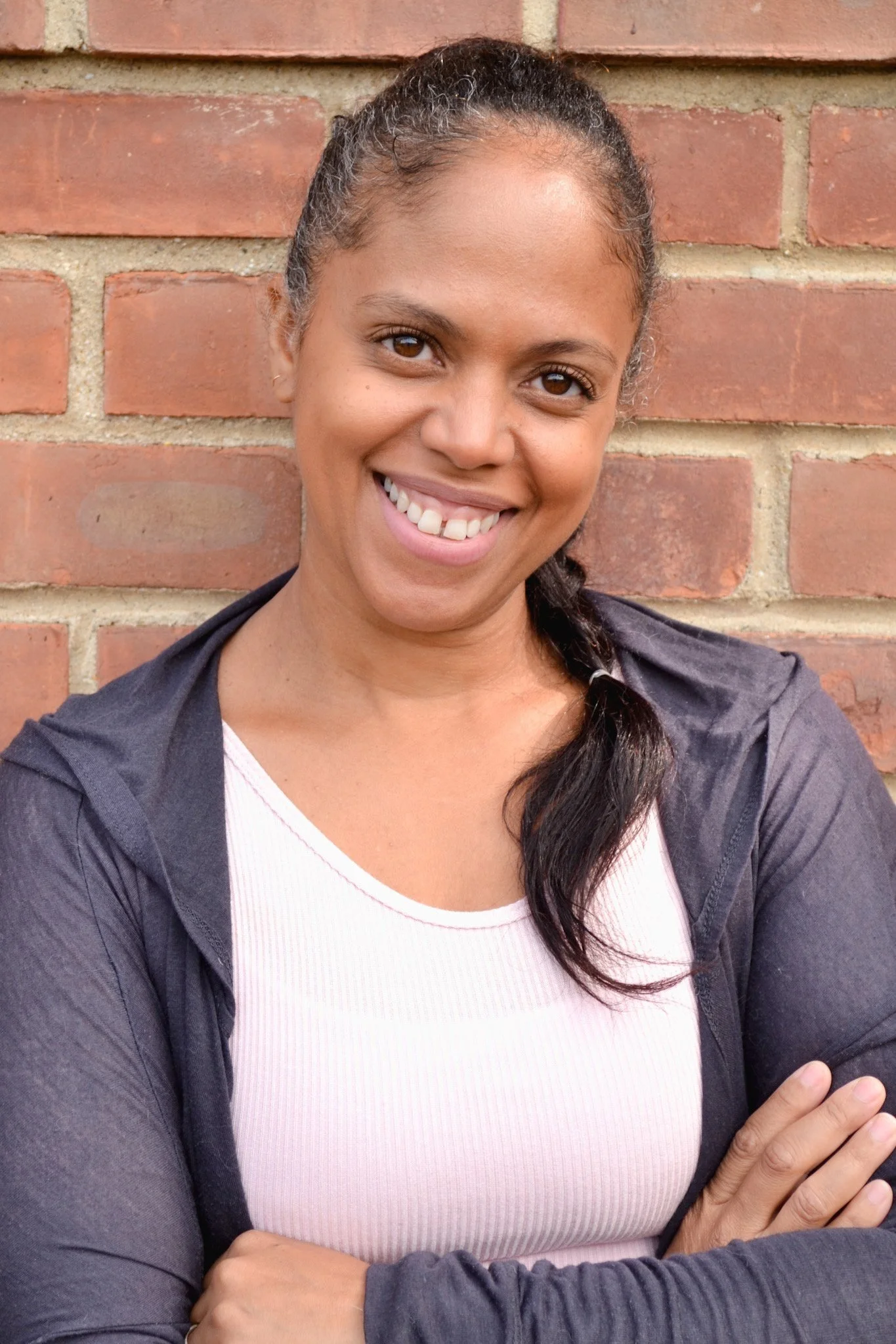 A woman smiling with arms crossed, standing in front of a brick wall.