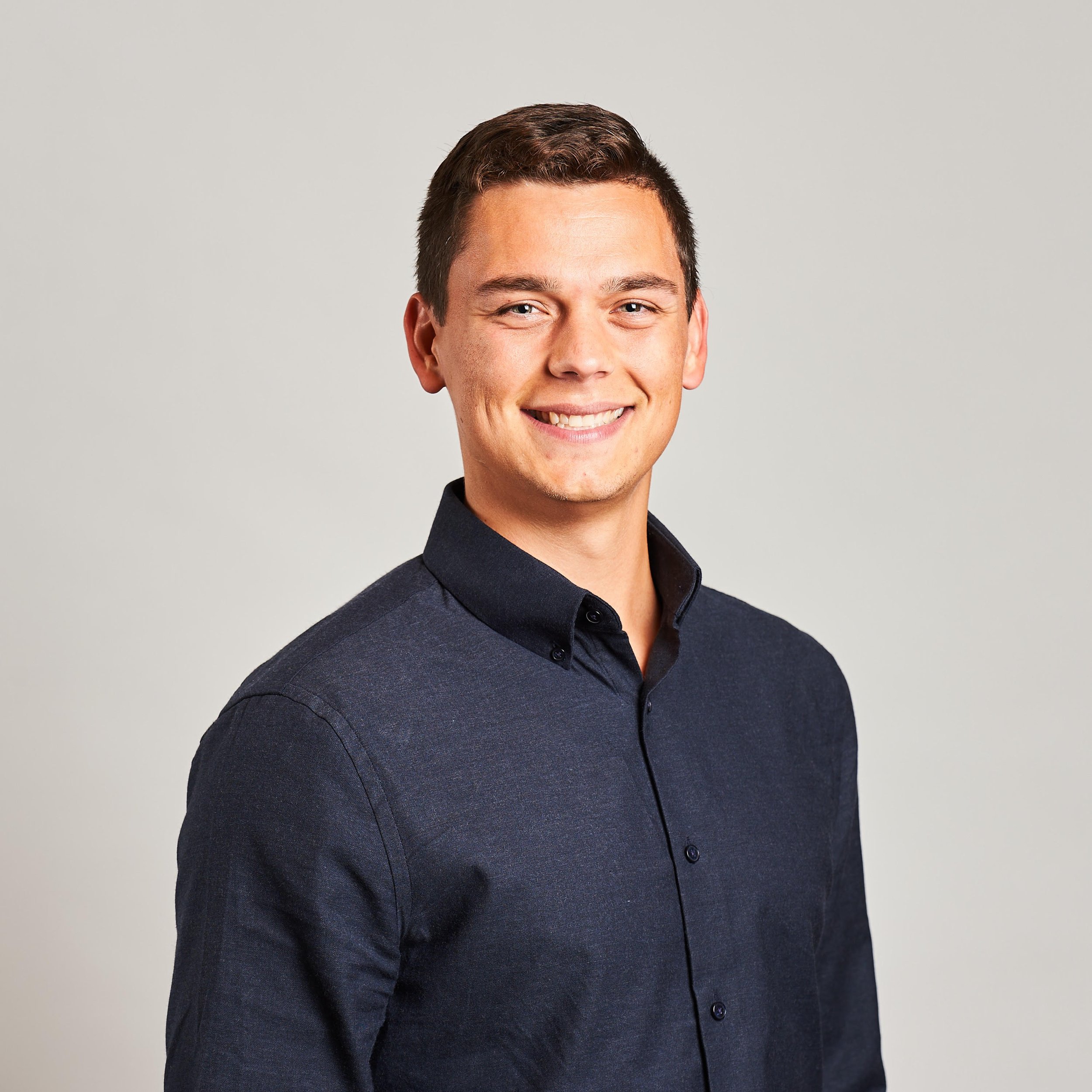 Portrait of a young man with short brown hair, smiling, wearing a dark button-up shirt, against a plain light background.