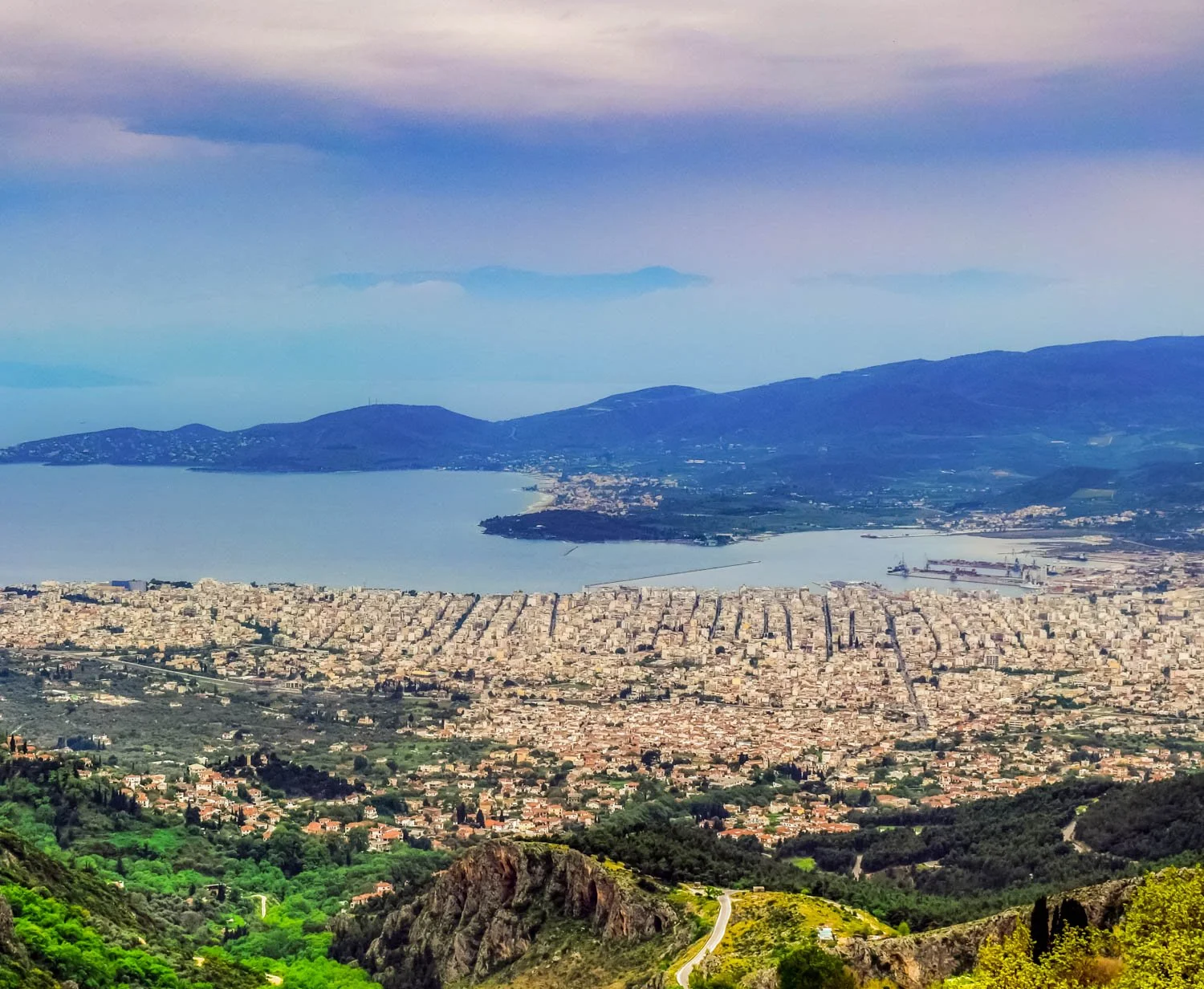 A panoramic view of a coastal city with a bay, surrounded by hills and mountains, featuring dense urban areas, green vegetation, and shipping docks.