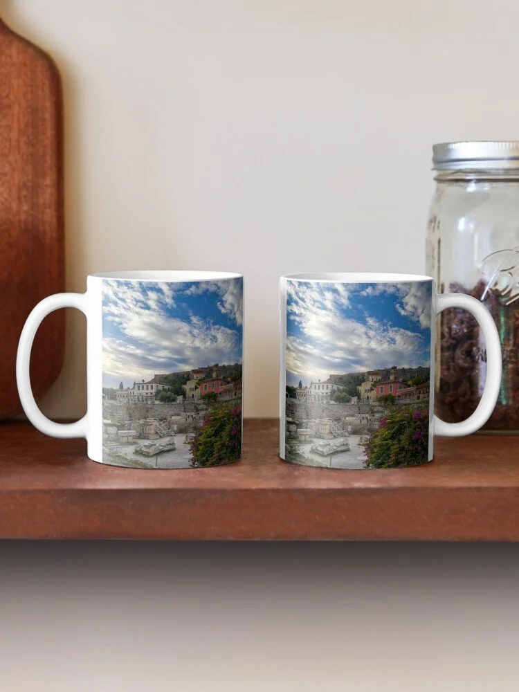 Two ceramic coffee mugs on a wooden shelf, each featuring a scenic cityscape with a cloudy sky, buildings, and trees.