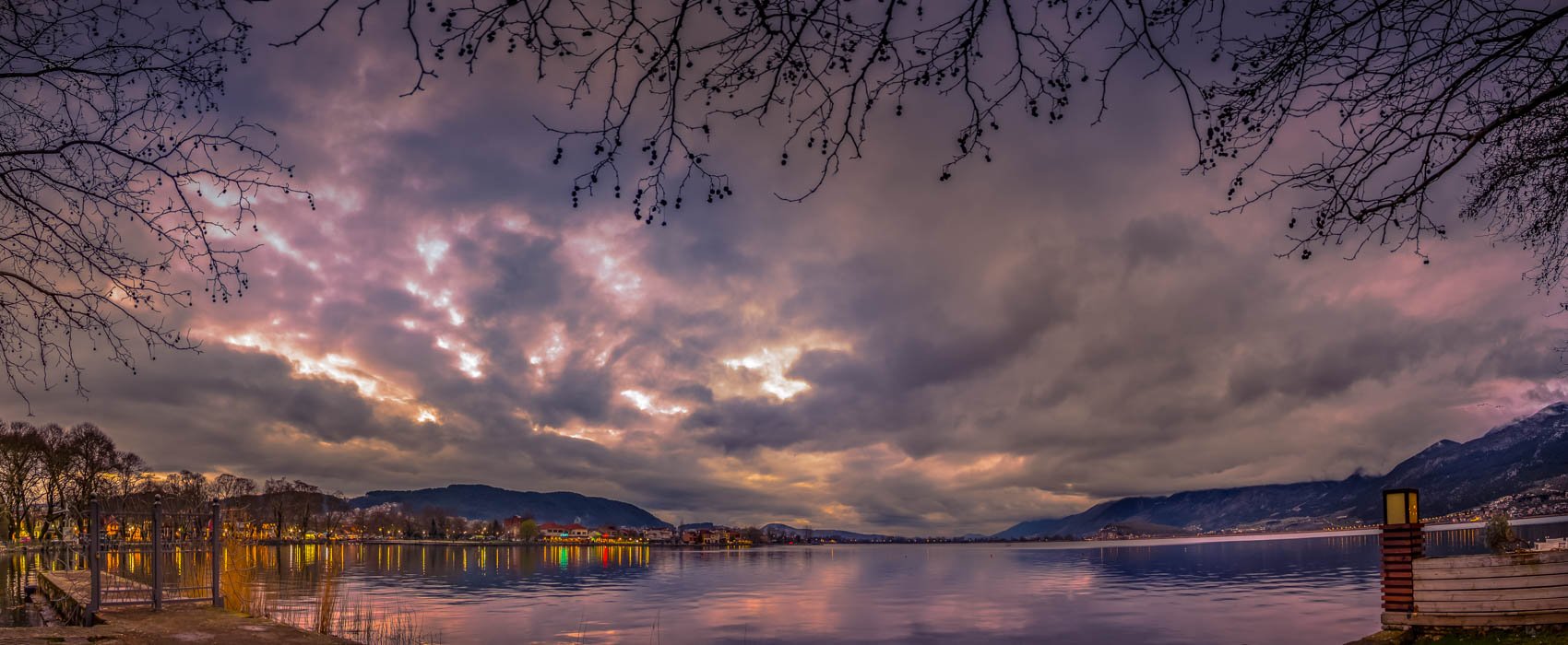 A body of water with a mountain range in the distance, a cloudy sky at sunset, and trees with bare branches in the foreground.
