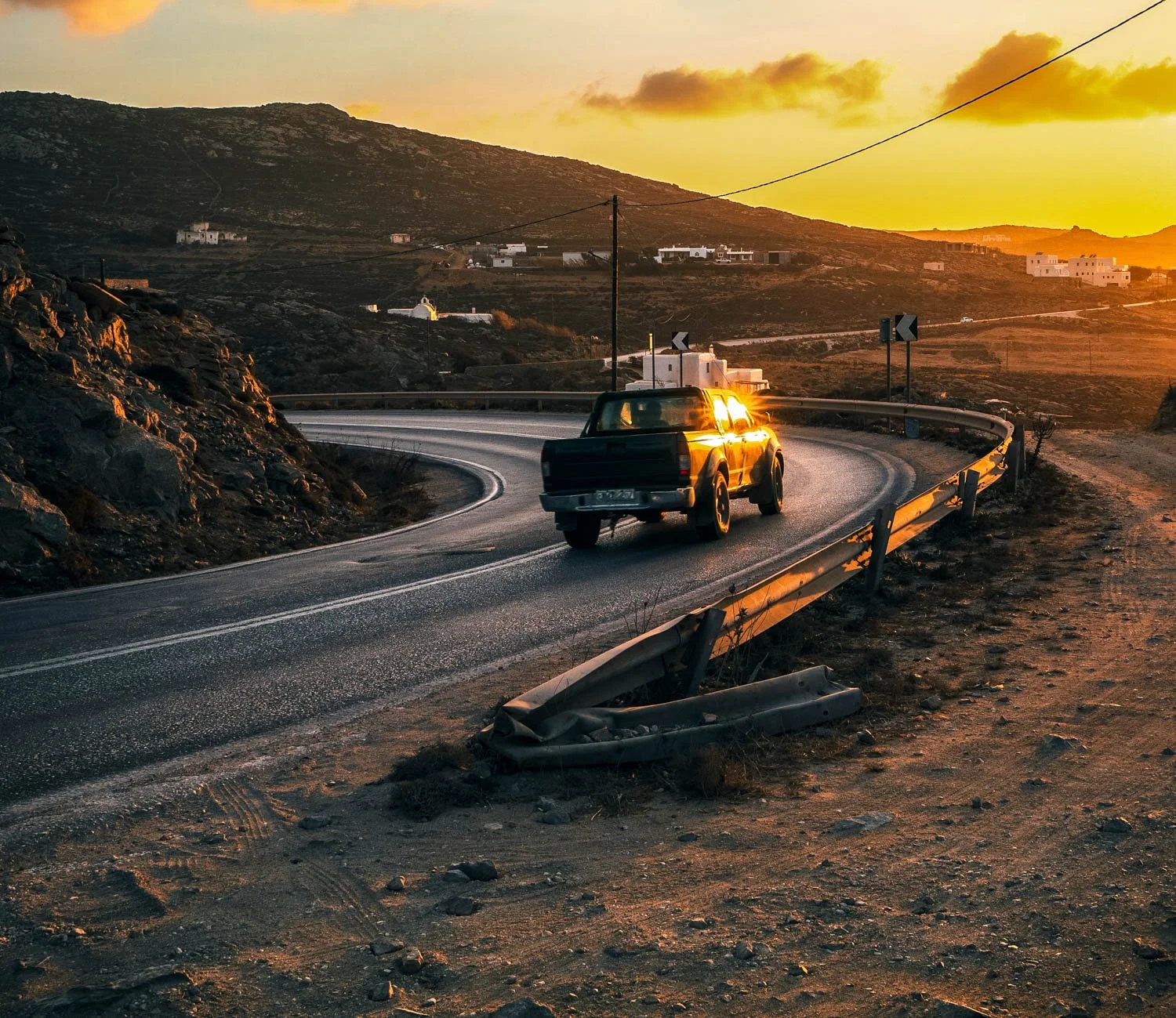 A black pickup truck drives on a winding mountain road during sunset, with a metal guardrail and rocky terrain on the side, and houses and hills in the background.