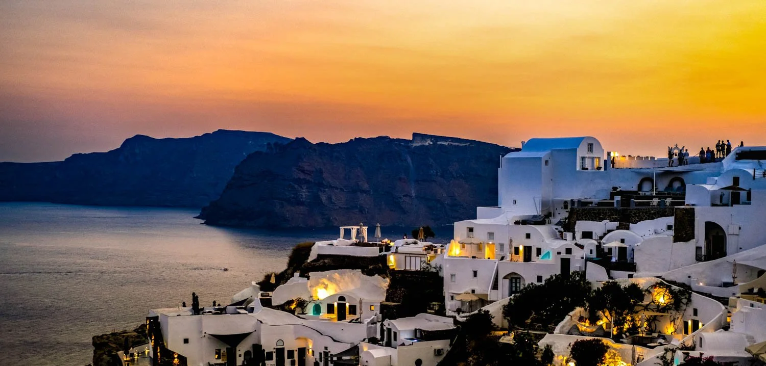 Sunset over white-washed buildings on a hillside in Santorini with the ocean and cliffs in the background.