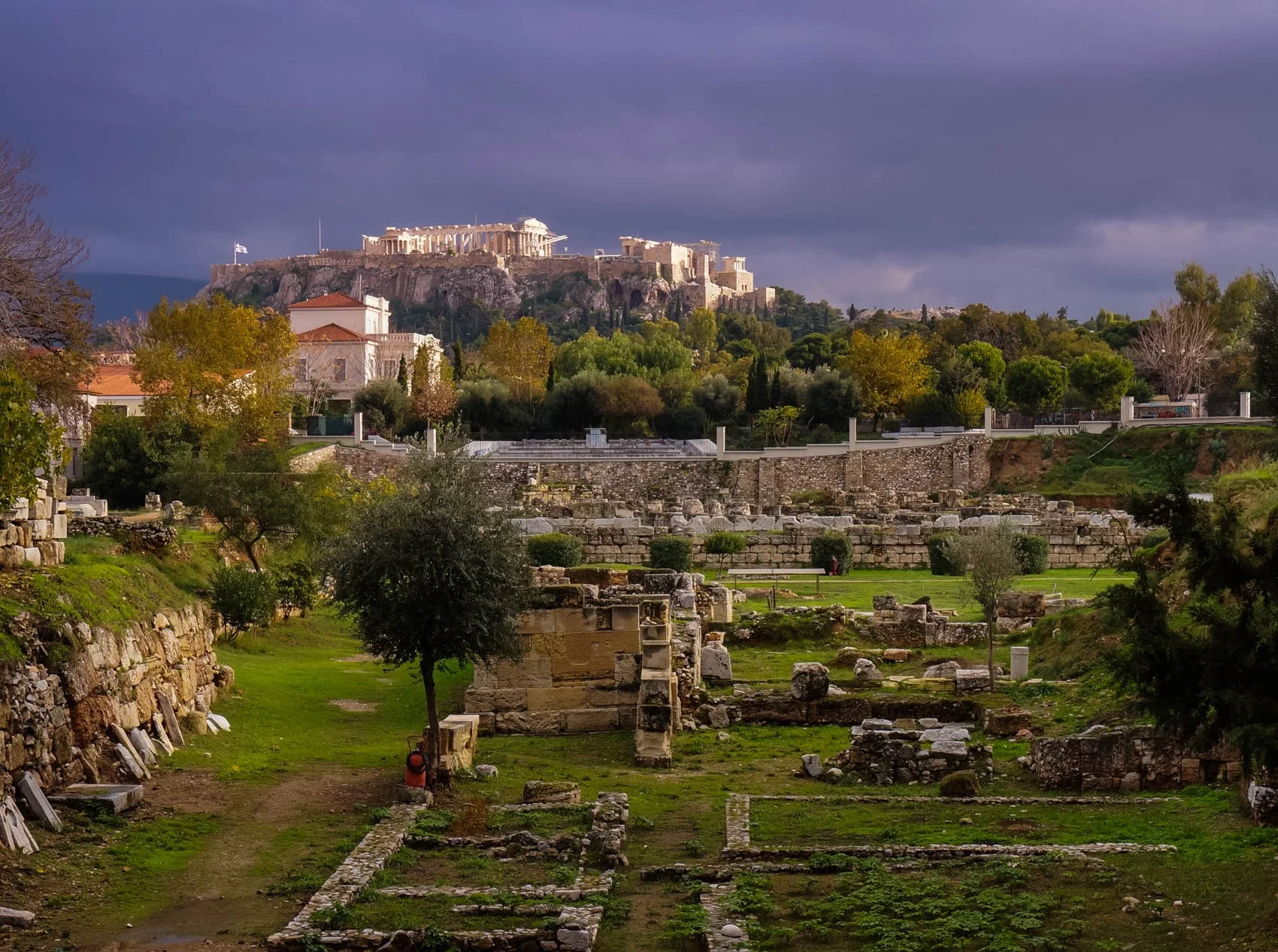 Ancient ruins with stone structures and greenery in foreground, trees and bushes, and the Parthenon atop the Acropolis in the background under a cloudy sky.