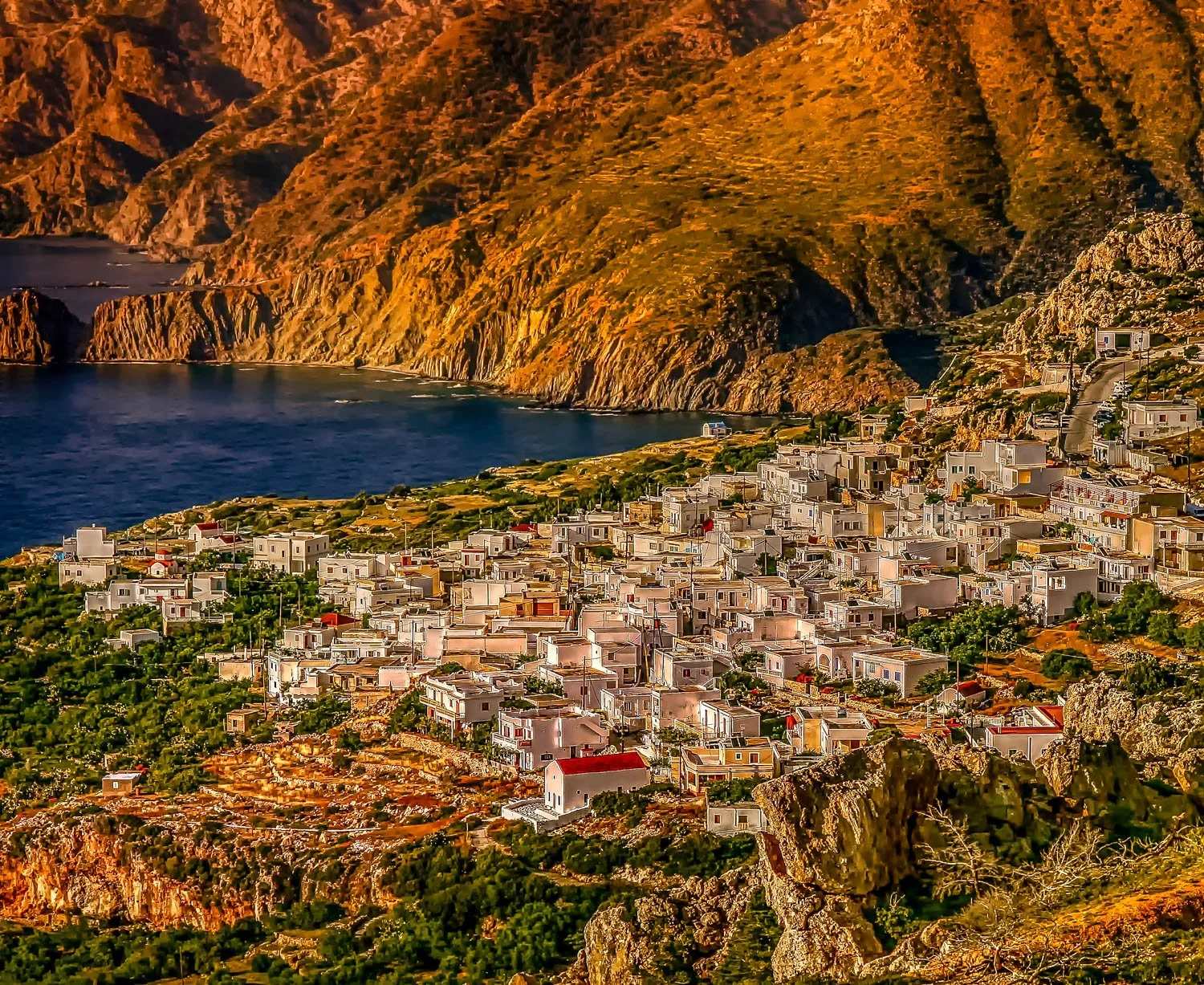 A coastal village with white buildings on a hillside overlooking the sea, with rugged mountains in the background.