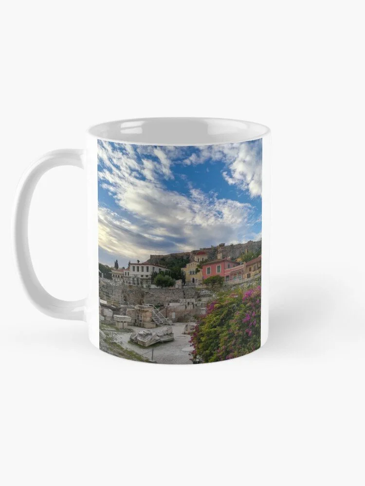A white mug with a scenic print of a hillside town under a partly cloudy sky, featuring colorful buildings and rocky ruins in the foreground.