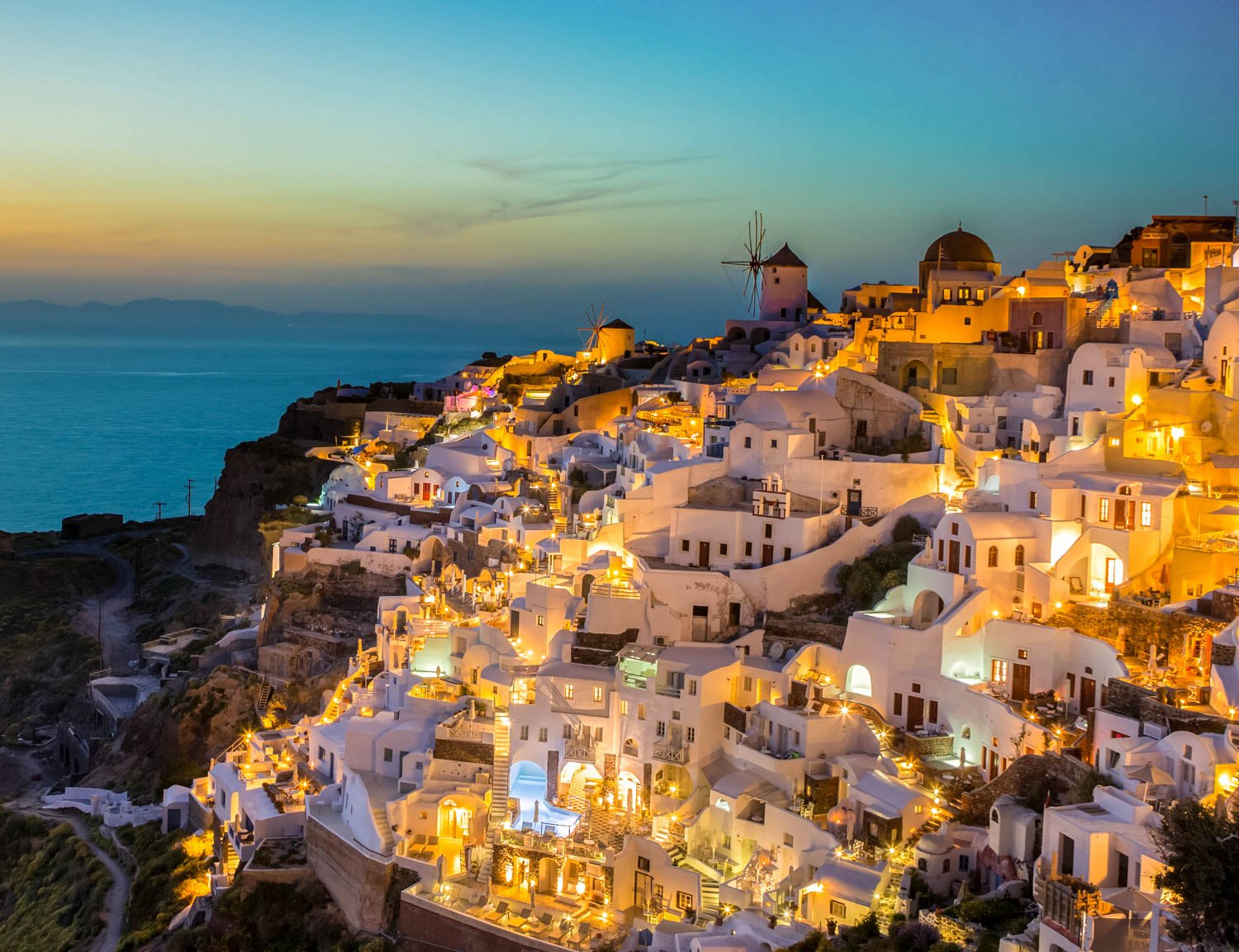 Scenic view of Santorini at dusk with white buildings illuminated along the hillside overlooking the sea, and windmills in the background.
