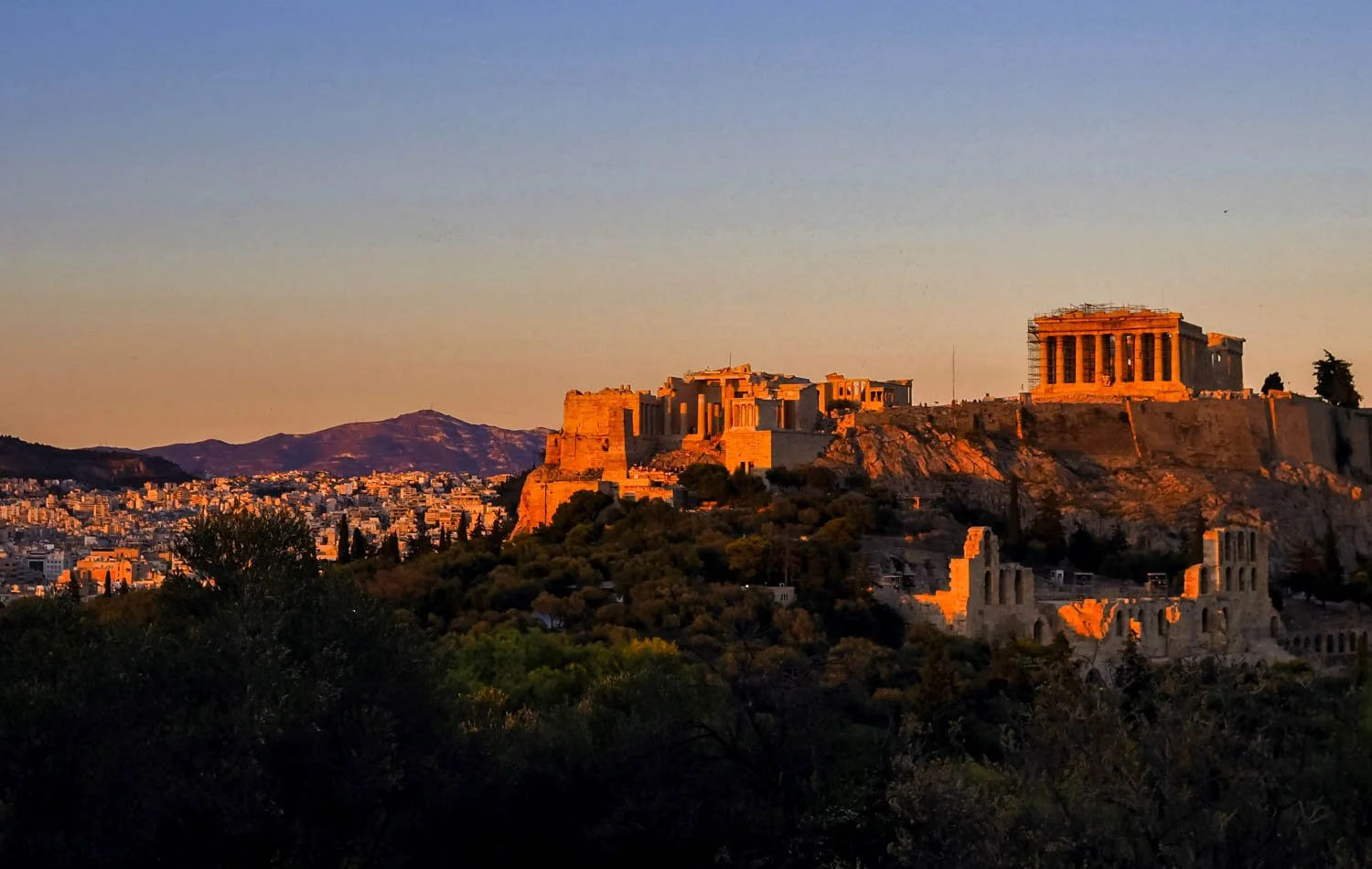 Sunset view of the Acropolis in Athens, Greece, with the Parthenon illuminated by the setting sun, overlooking the city and mountains.