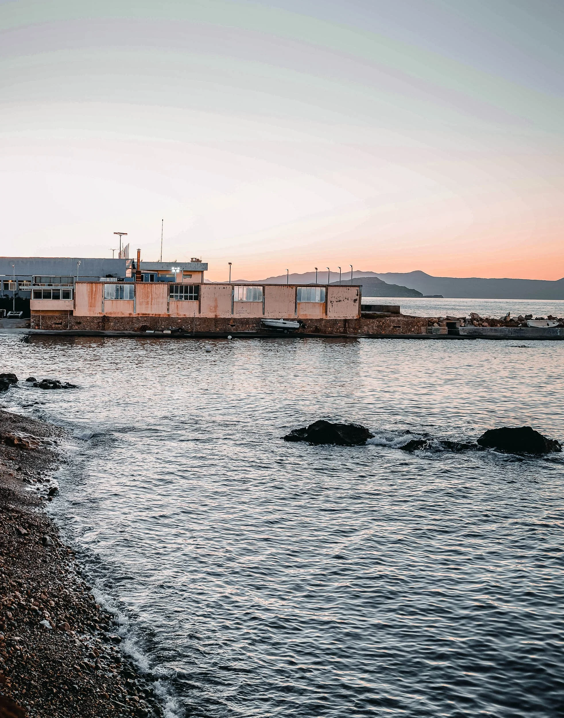 Sunset over a harbor with a building on the water, rocks in the foreground, and mountains in the distance.