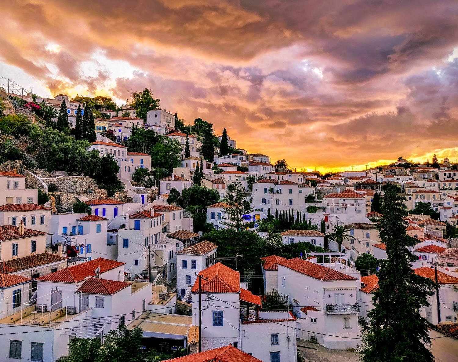 A hillside town with white buildings and red-tiled roofs at sunset, under a colorful cloud-filled sky.
