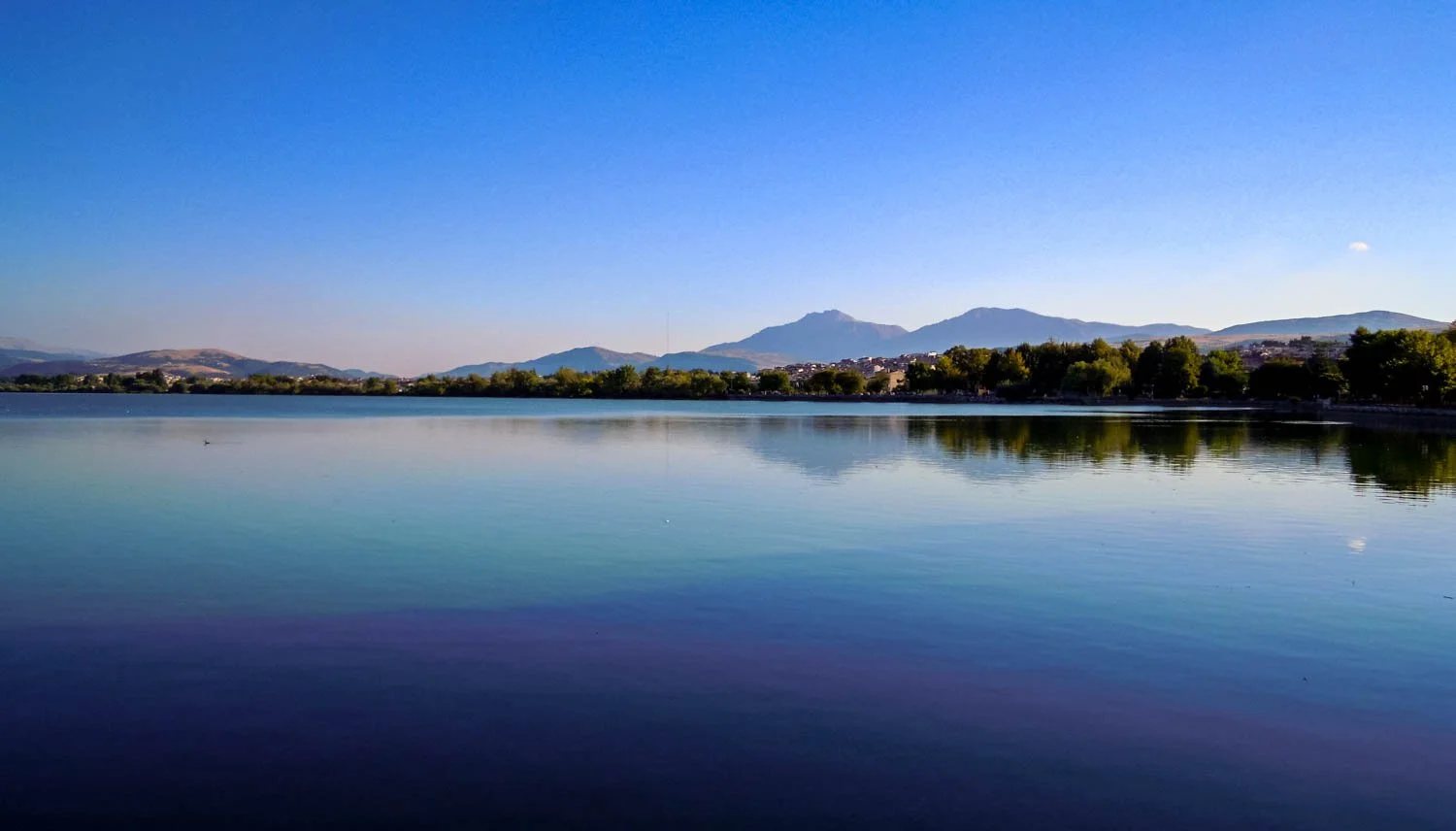 Calm lake reflecting surrounding trees and distant mountains under a clear blue sky.