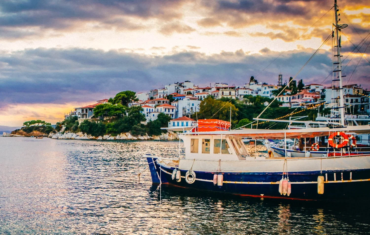 Boats anchored in a calm harbor with a hillside town of white buildings and red-tiled roofs, under a cloudy sky during sunset.