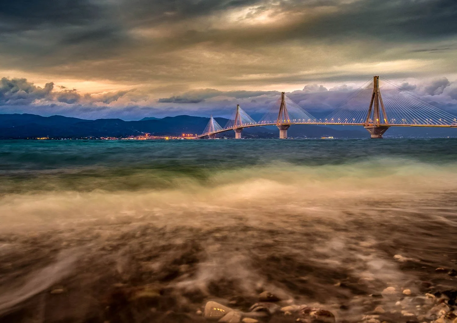 A large bridge with multiple towers and cables extending across a body of water during dusk, with mountains and clouds in the background.