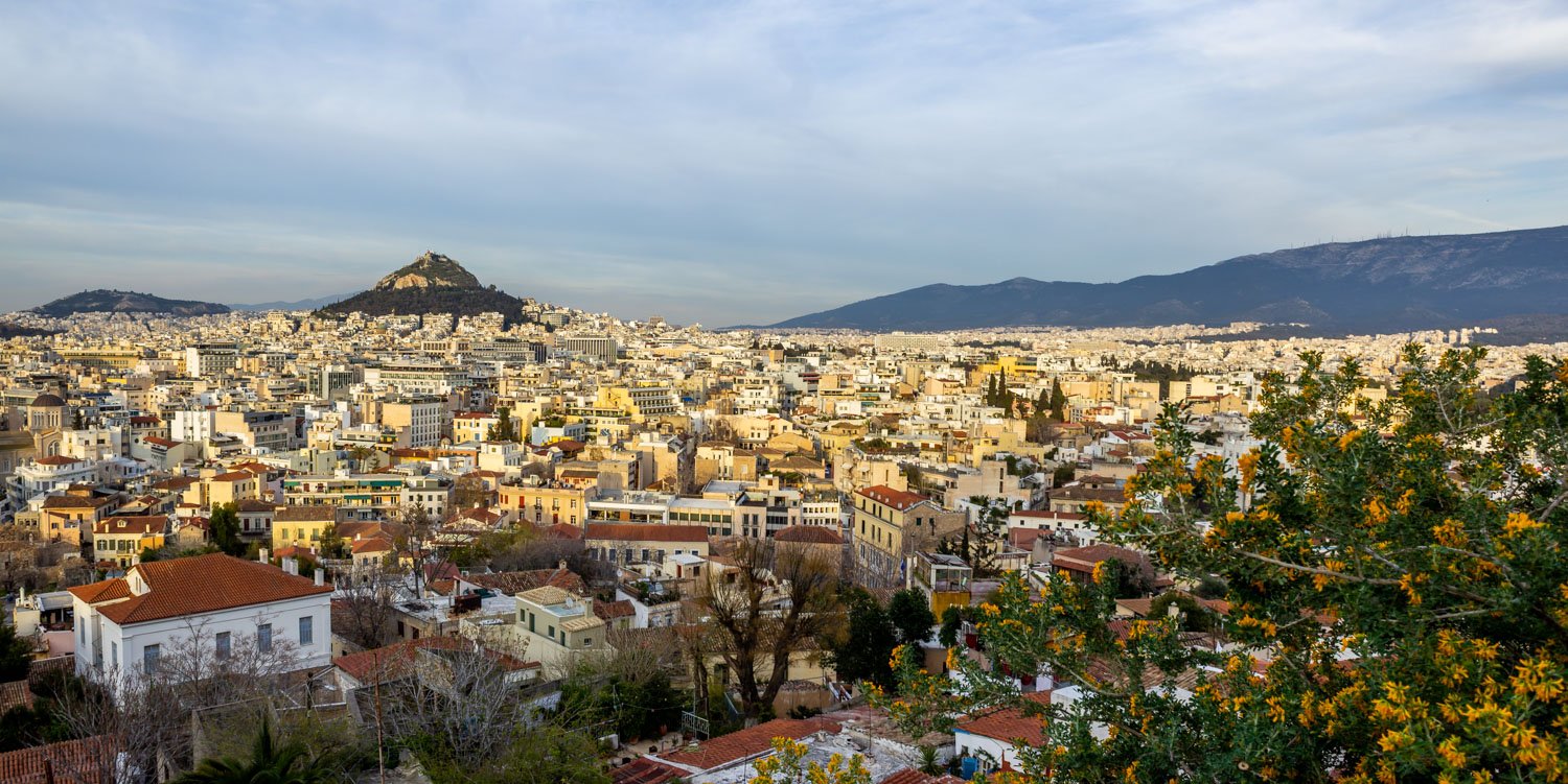 Panoramic view of Athens, Greece, with white and beige buildings, Mount Lycabettus in the background and a flowering orange tree in the foreground.