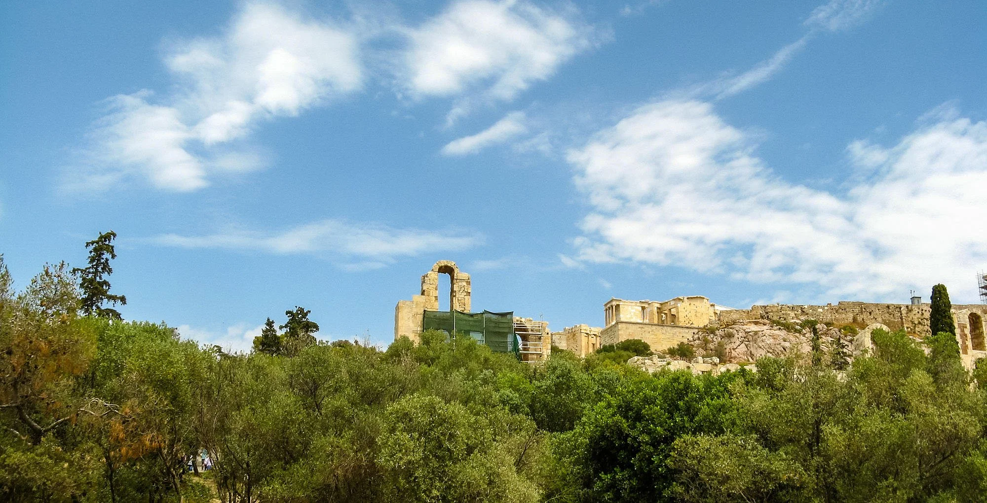 Ancient ruins on a hilltop with a partially standing arch, surrounded by green trees and a blue sky with scattered clouds.