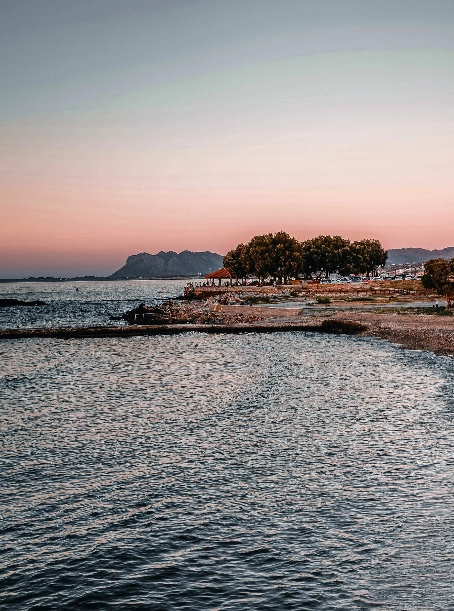 A calm seaside scene at sunset with a small pier, trees, mountains in the background, and a clear sky with pastel colors.