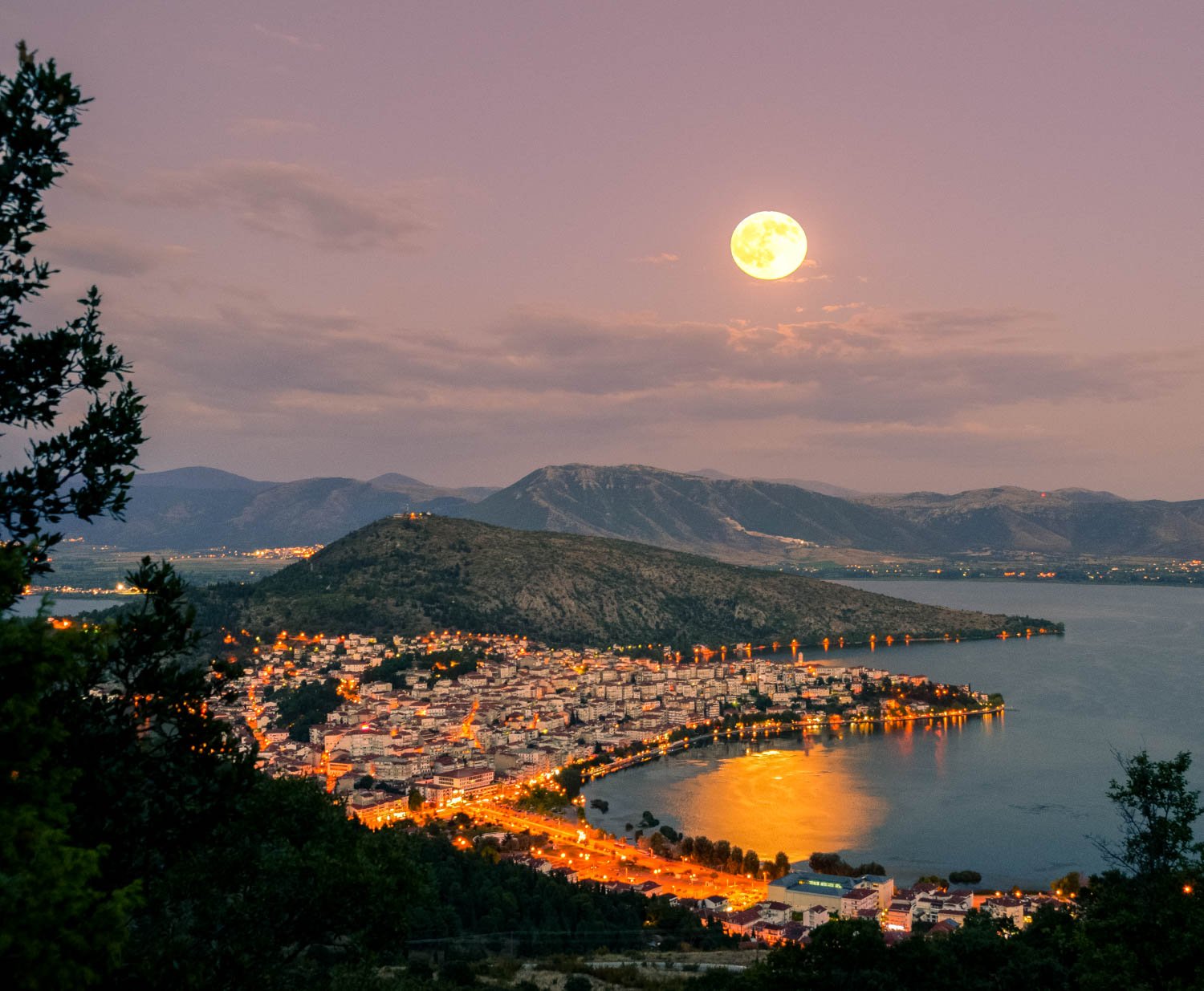 Nighttime landscape of a town between a lake and hills, with a large, bright full moon in a pink and purple sky reflecting off the water.
