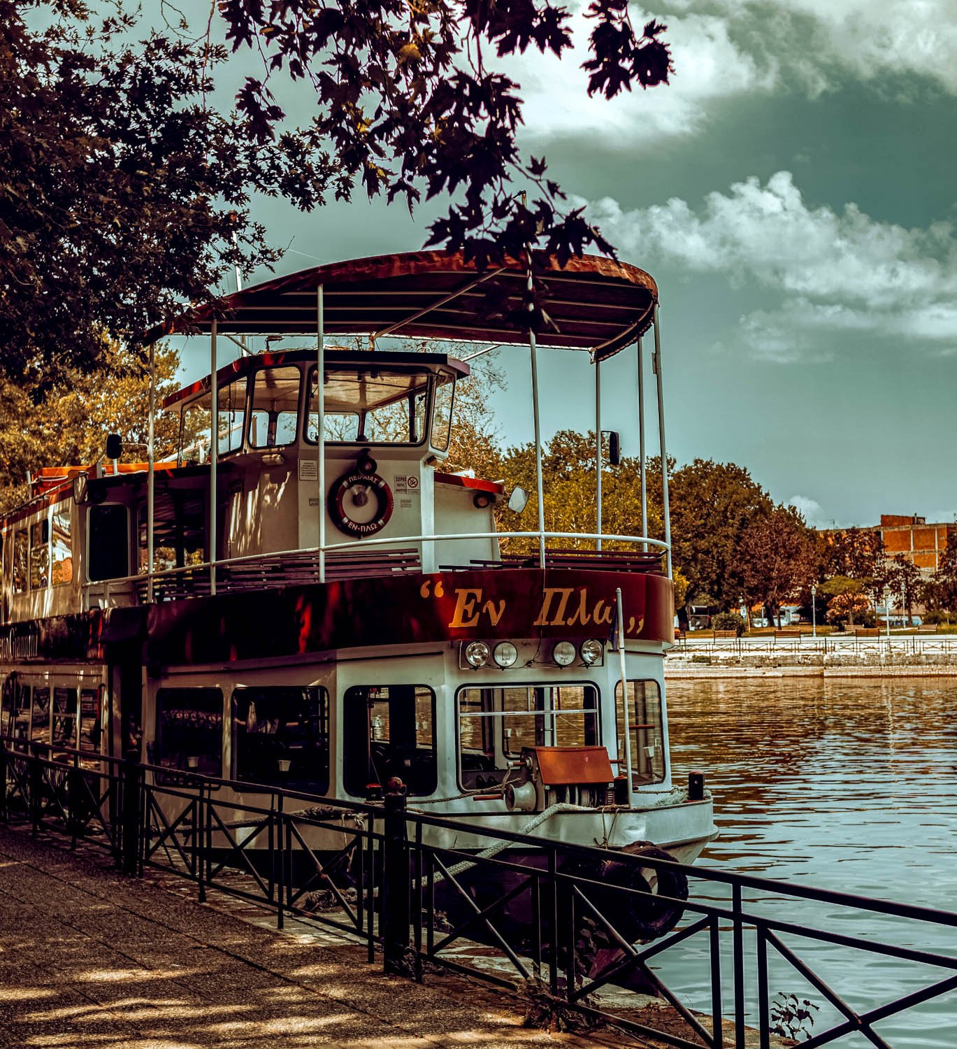 A two-level boat docked along a waterfront promenade with an iron railing. The boat has a red and white color scheme with Greek writing on the front. Trees with foliage and a partly cloudy sky are visible in the background.