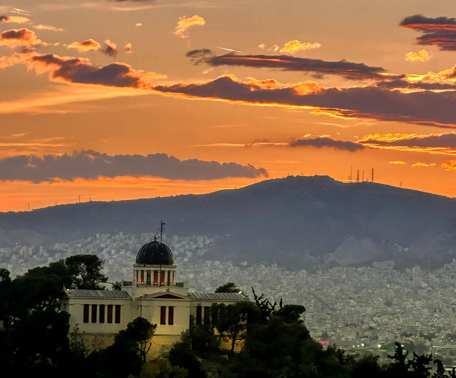 A sunset over a city with a hill in the background. In the foreground, there is a historic building with a dome and columns surrounded by trees.