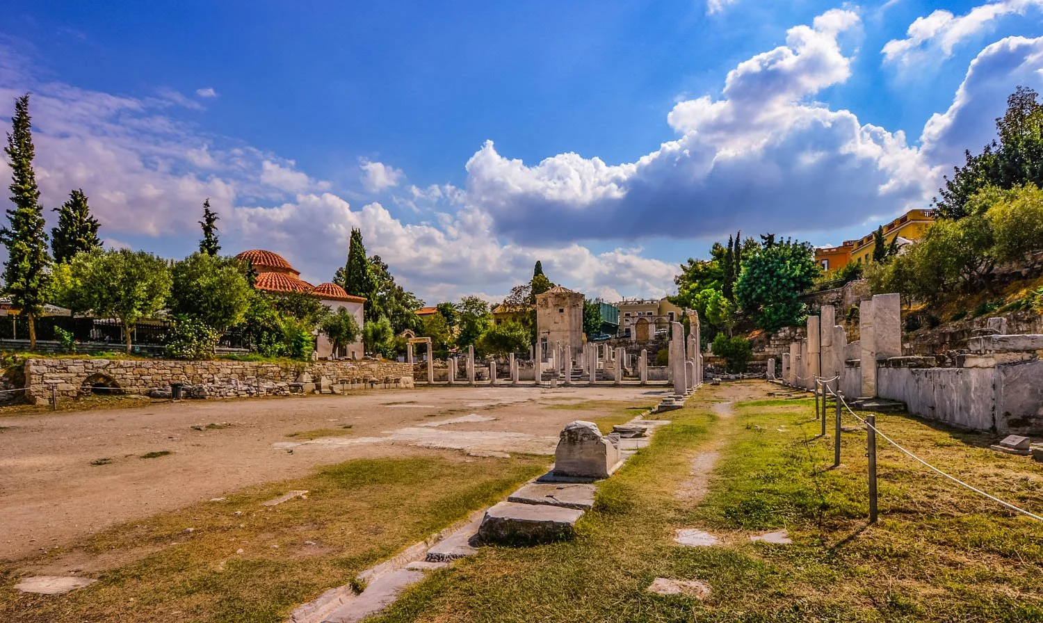 Ancient ruins with stone columns and walls, green trees, colorful houses, and a partly cloudy sky.