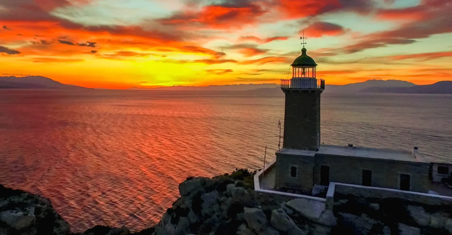 Lighthouse on a rocky coastline during a colorful sunset with orange, yellow, and pink clouds over the ocean and distant mountains.