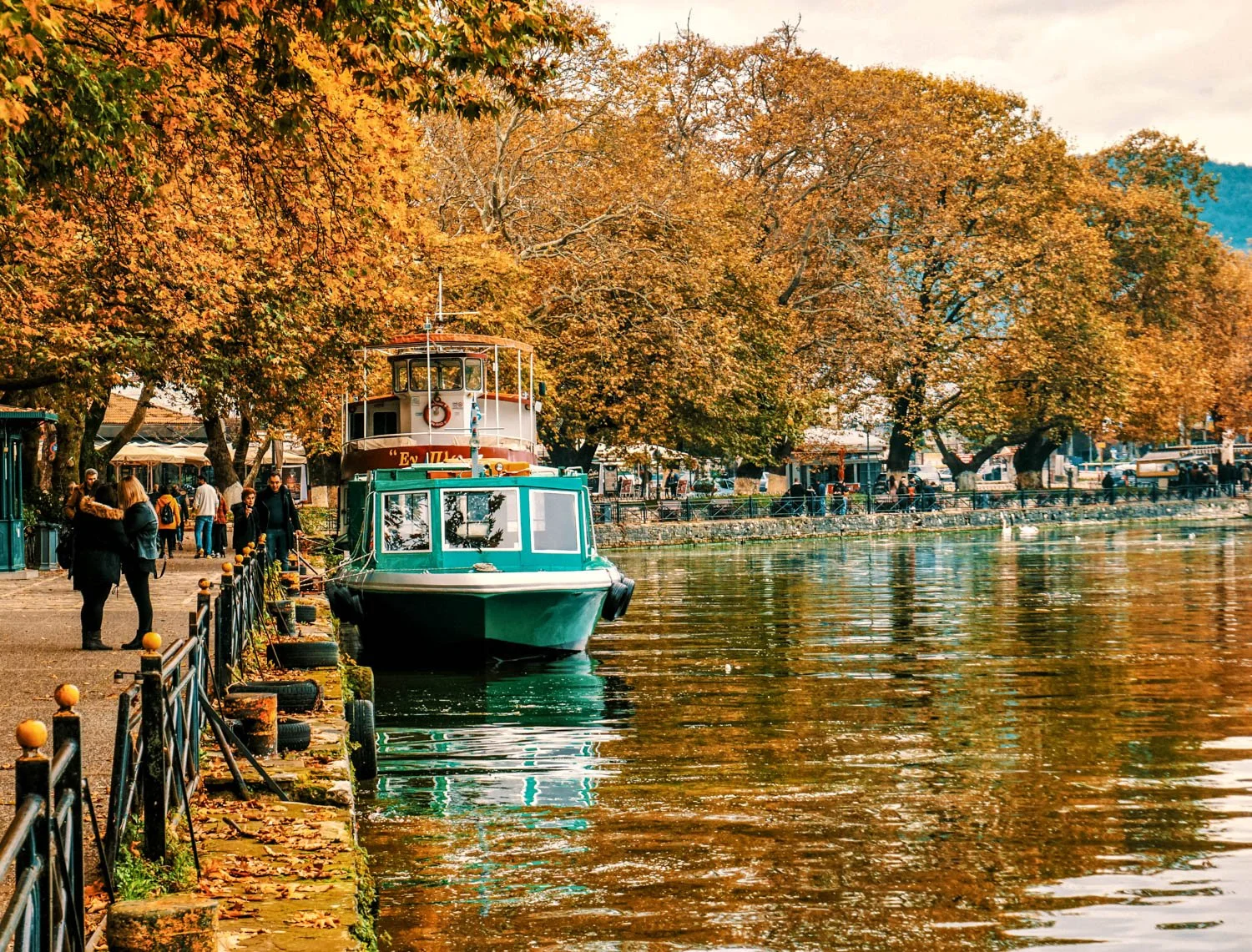 Boats docked along a riverbank with trees displaying fall foliage and people walking nearby.