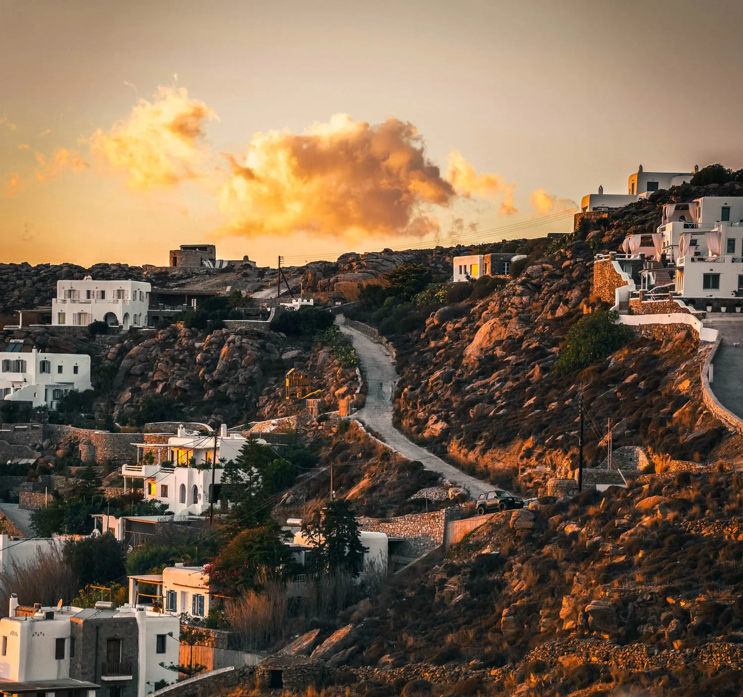 Sunset over a hillside with white Mediterranean-style houses, rocky terrain, and a winding road.