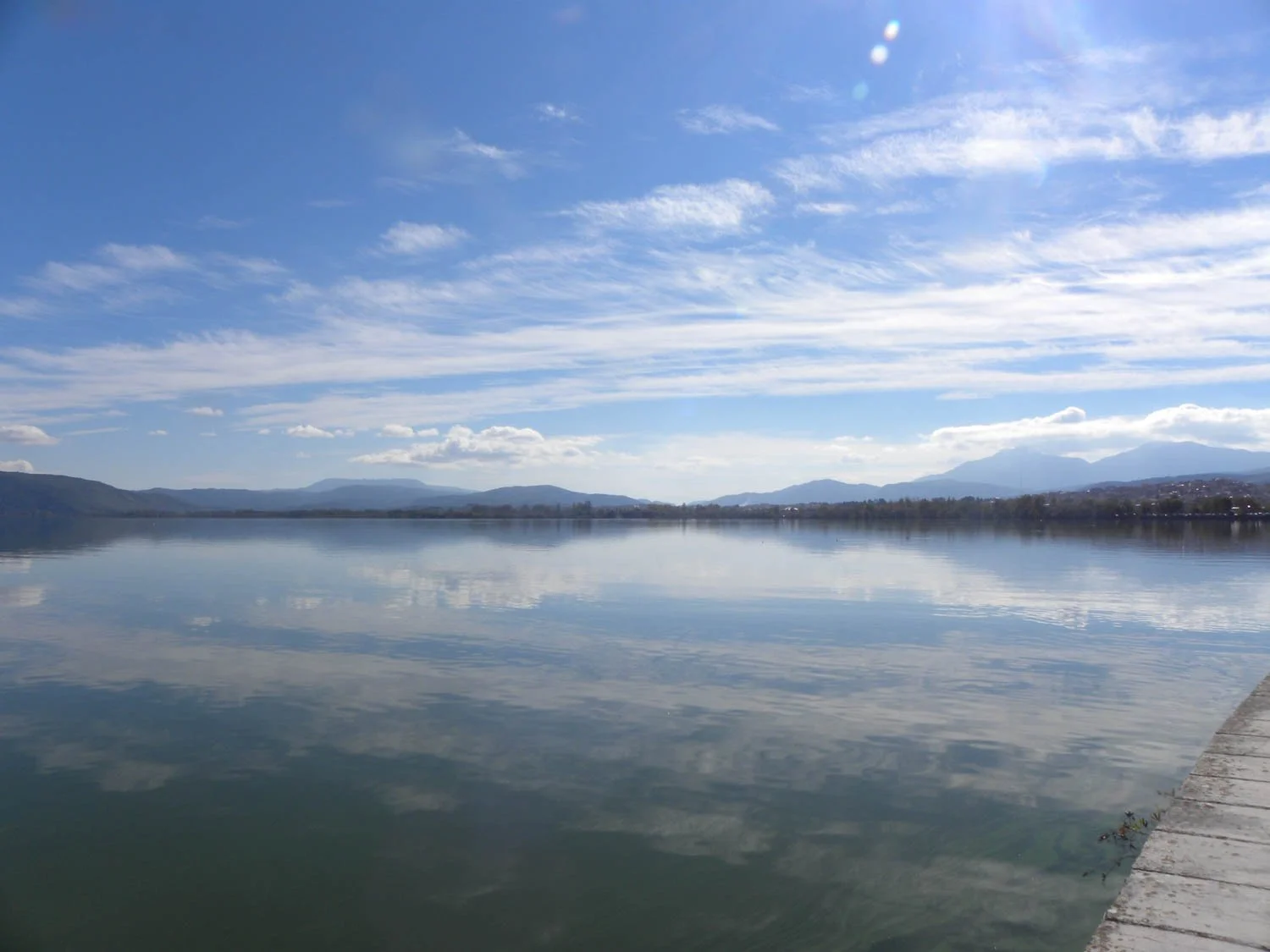 A calm lake reflecting a blue sky with scattered clouds, mountains in the distance, and a wooden dock on the right side.