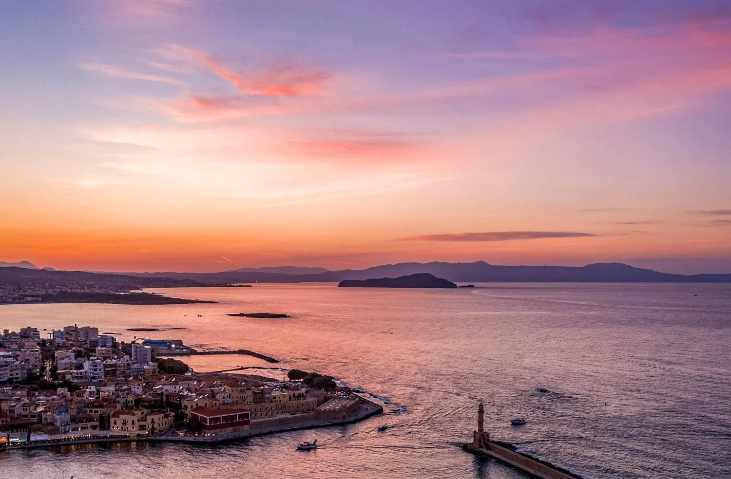 A scenic view of a coastal city during sunset with pink and purple sky, calm water, boats, and a lighthouse on a pier.