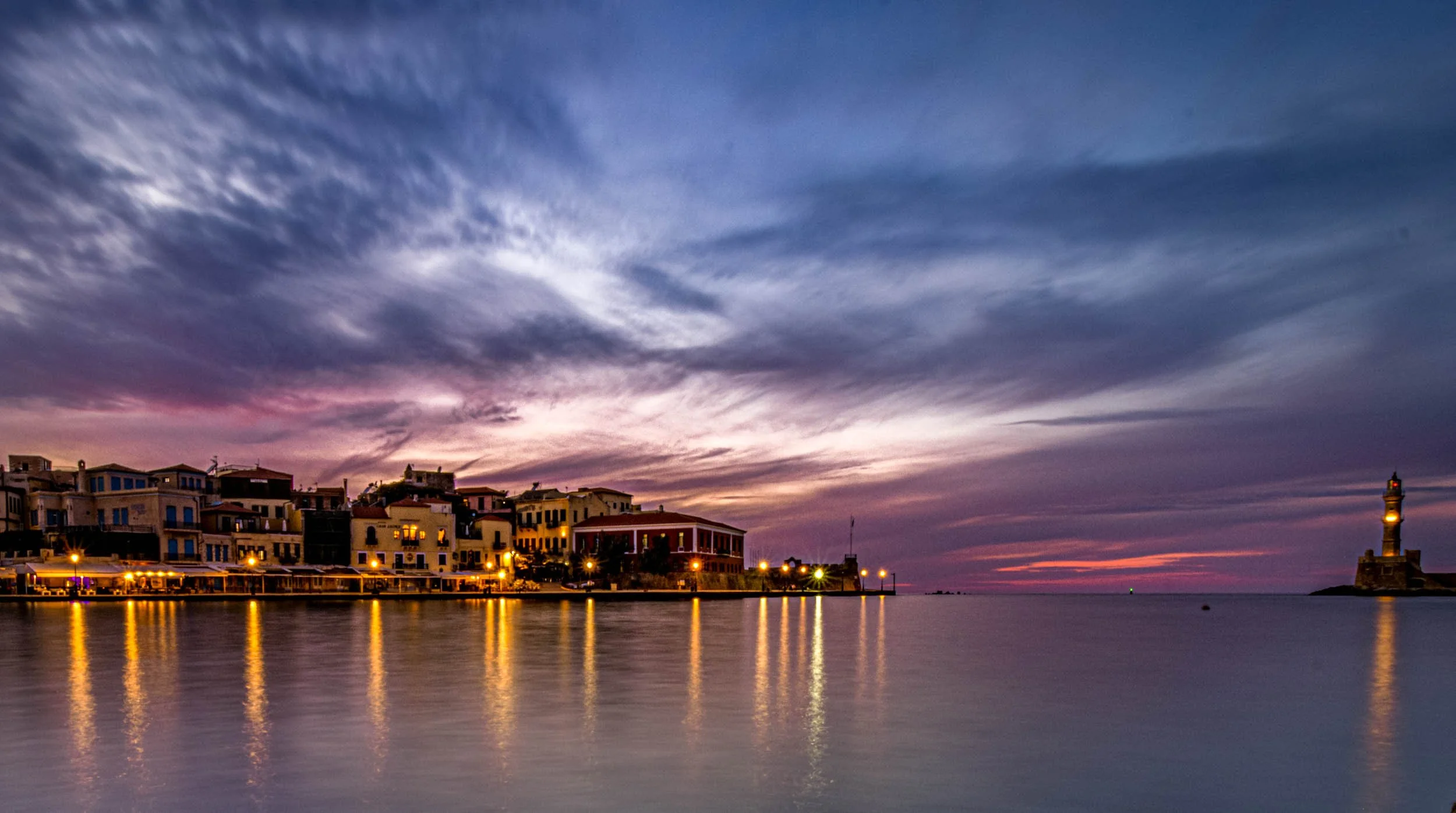 A scenic view of a harbor at sunset with colorful houses along the water and a lighthouse on a small island to the right, clouds in a vibrant sky reflecting on the calm water.