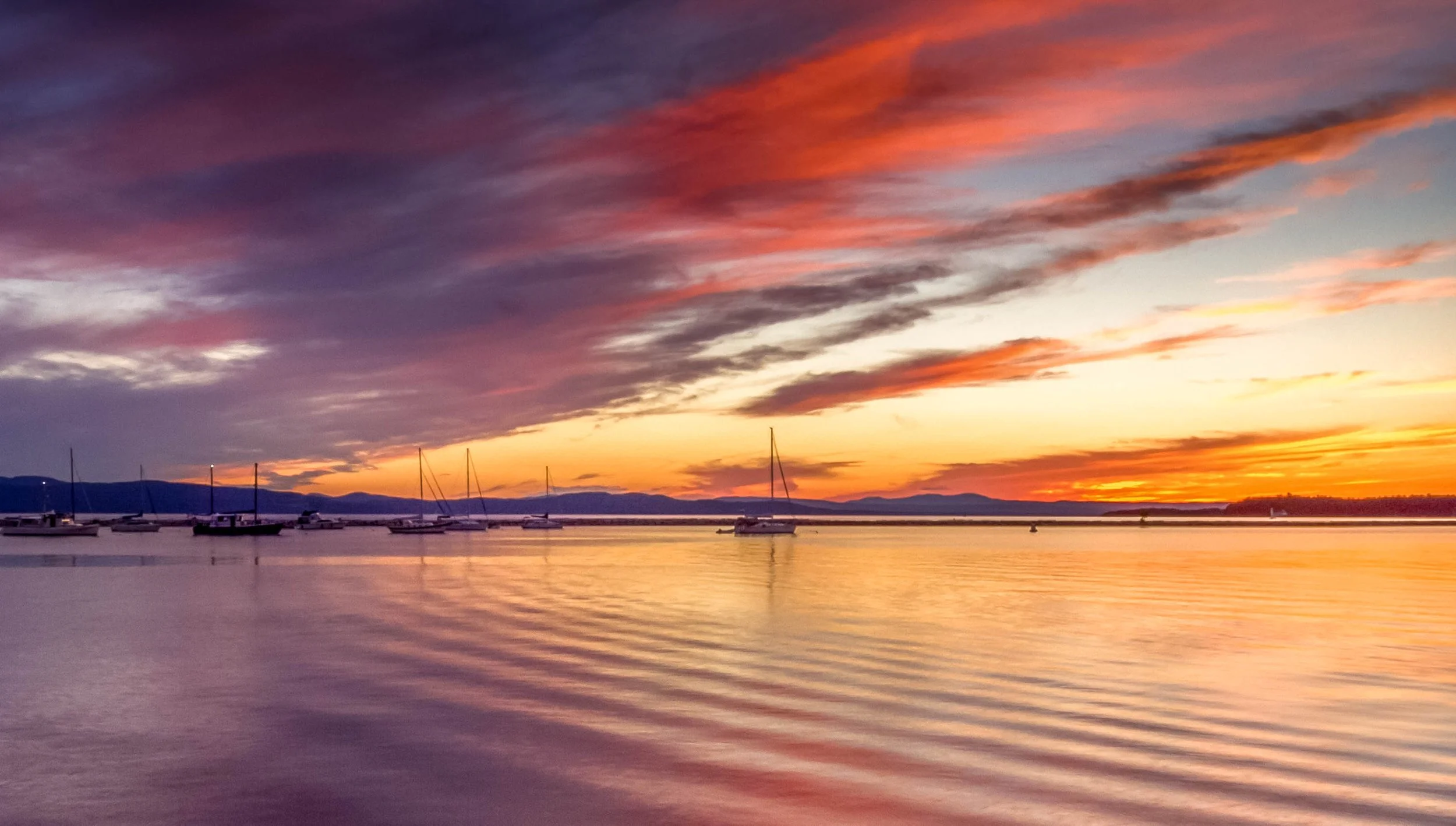 A scenic sunset over a calm body of water with sailing boats anchored in the distance and colorful clouds in the sky.