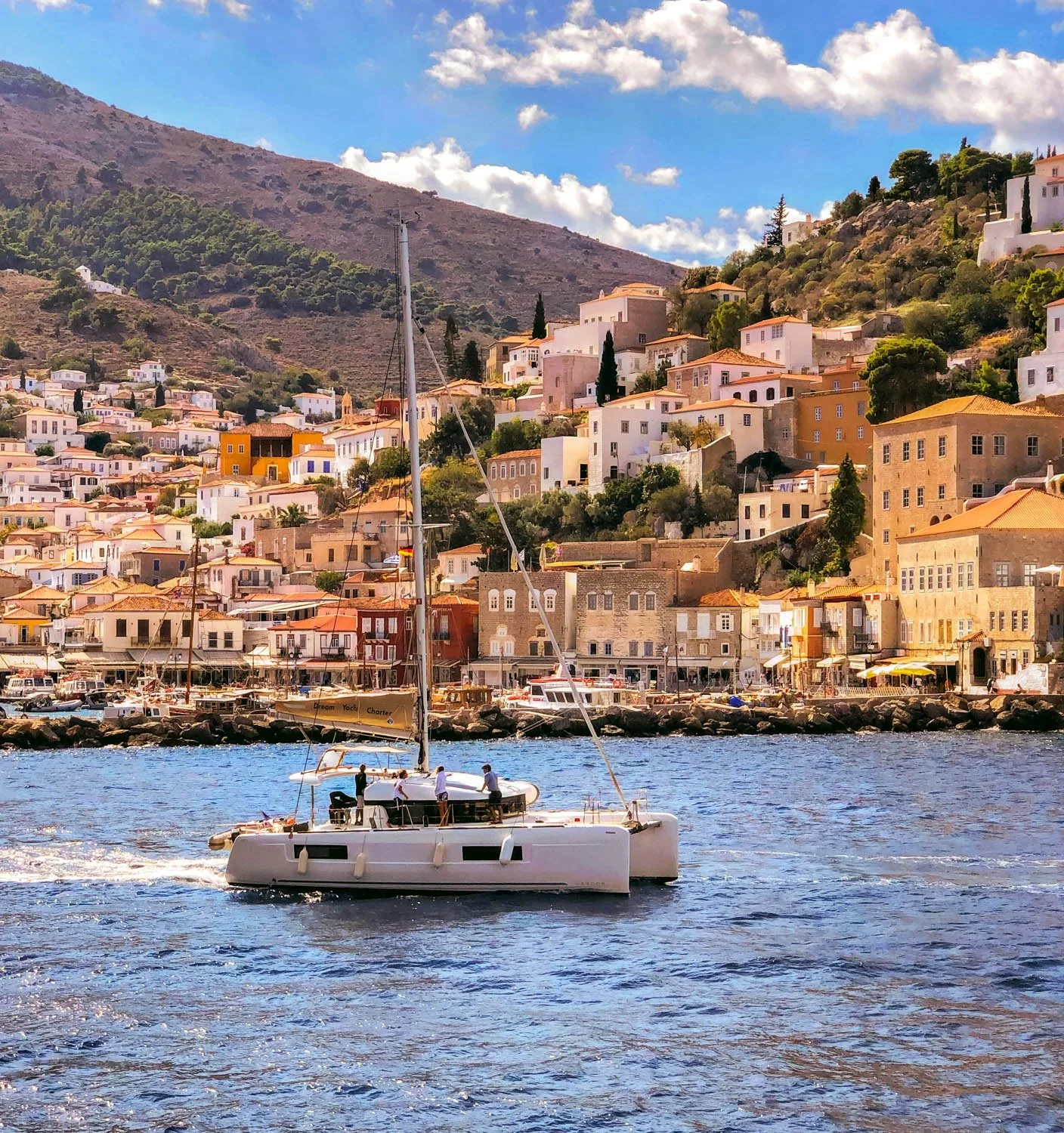 A white sailboat on the water with a hillside town in the background, featuring colorful buildings and green trees.