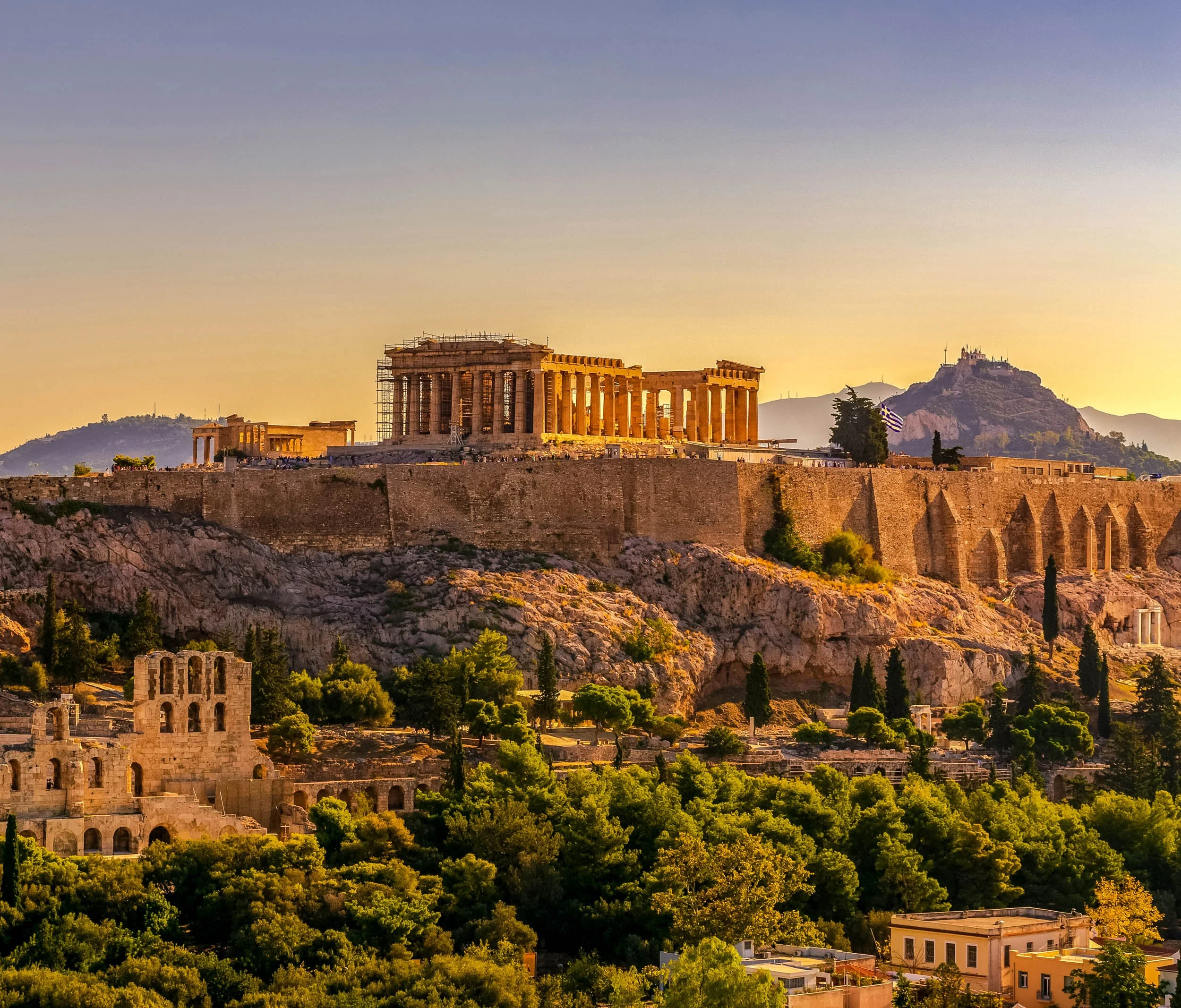 Sunset view of the Acropolis in Athens, Greece, featuring the Parthenon temple on a hilltop with surrounding ancient ruins, greenery, and mountains in the background.