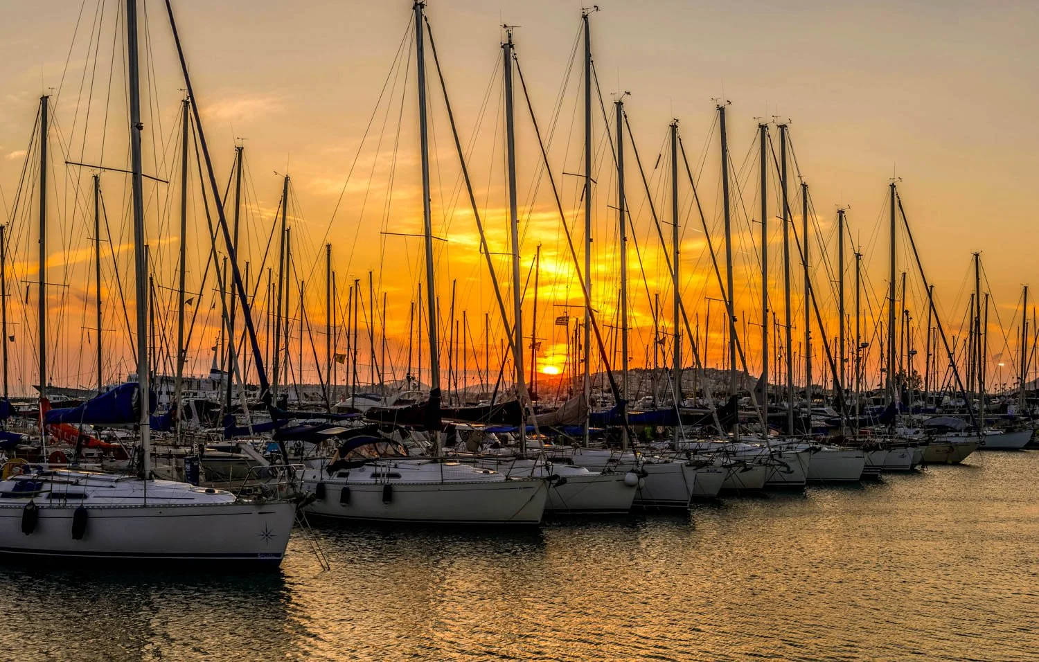 A marina with numerous sailboats docked at sunset, with the sky painted in warm orange and yellow hues.