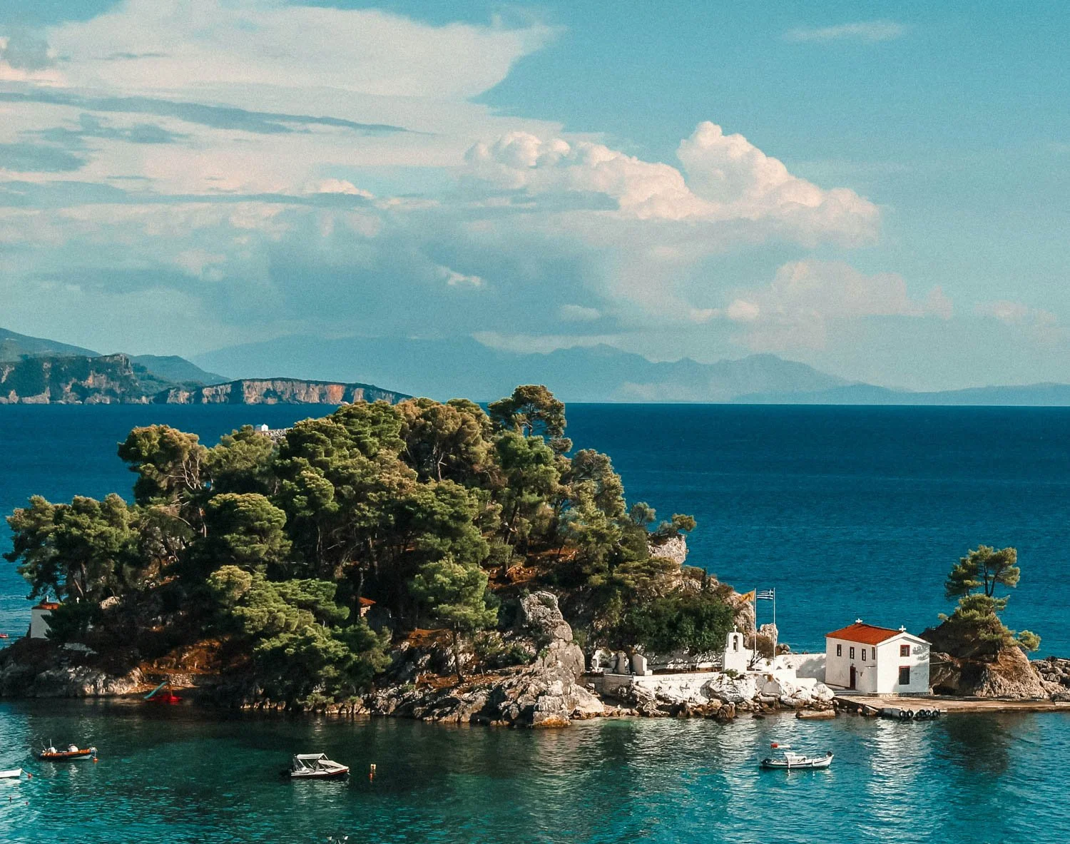 Scenic view of a small island with trees and white buildings, surrounded by blue waters, with boats floating nearby and mountains in the background under a partly cloudy sky.