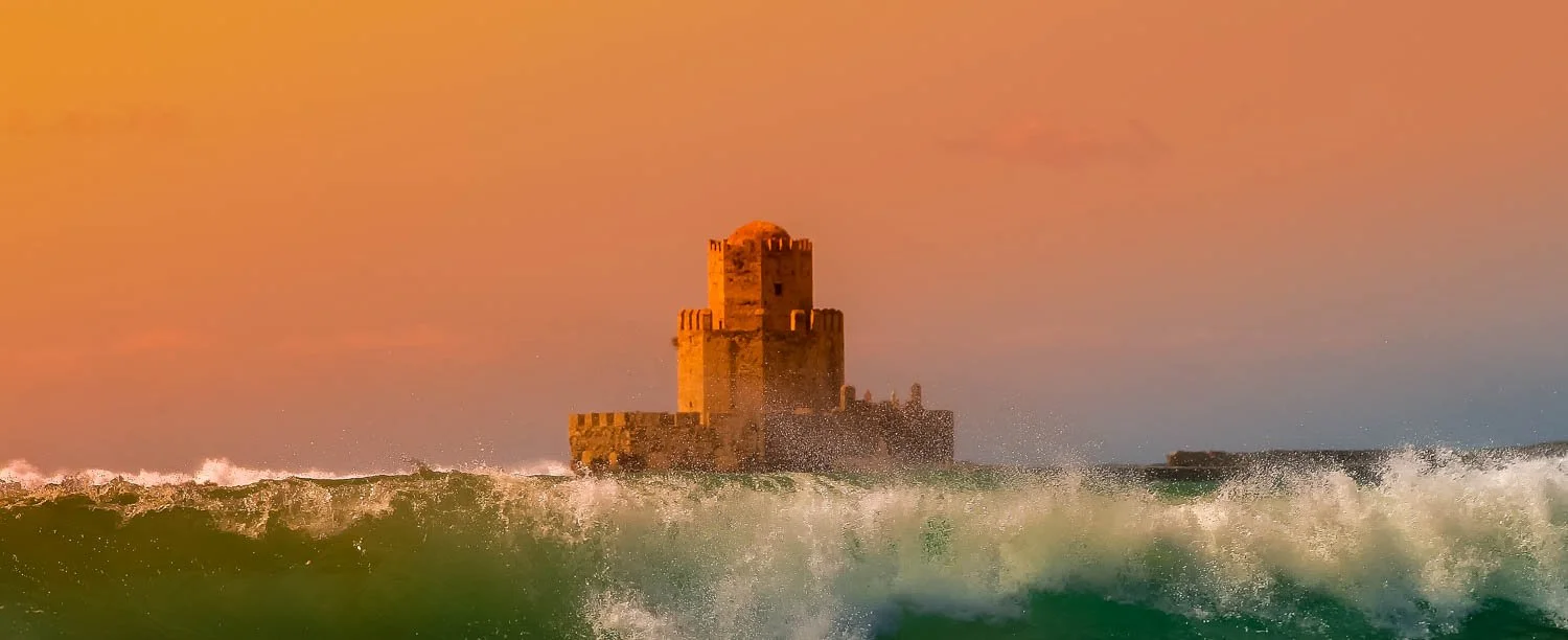 A historic stone castle with battlements stands in the ocean, partly obscured by large crashing waves during orange-hued sunset sky.
