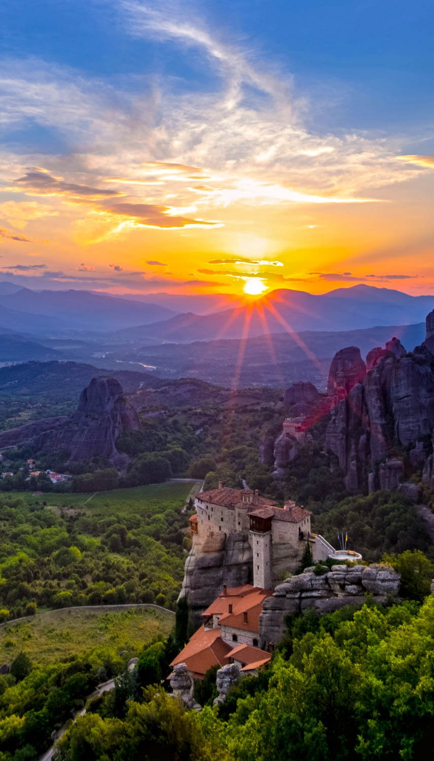 Sunset over mountainous landscape with rock formations and a monastery built on a rocky cliff, surrounded by green trees.