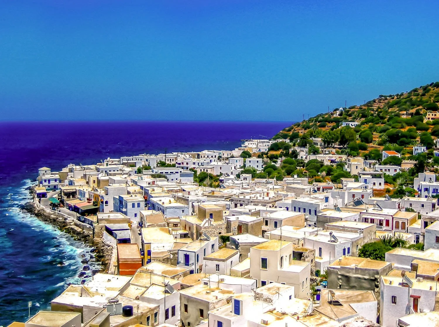 A coastal town with white buildings along the shore, turquoise and deep blue water, and a hilly landscape covered with green trees in the background.
