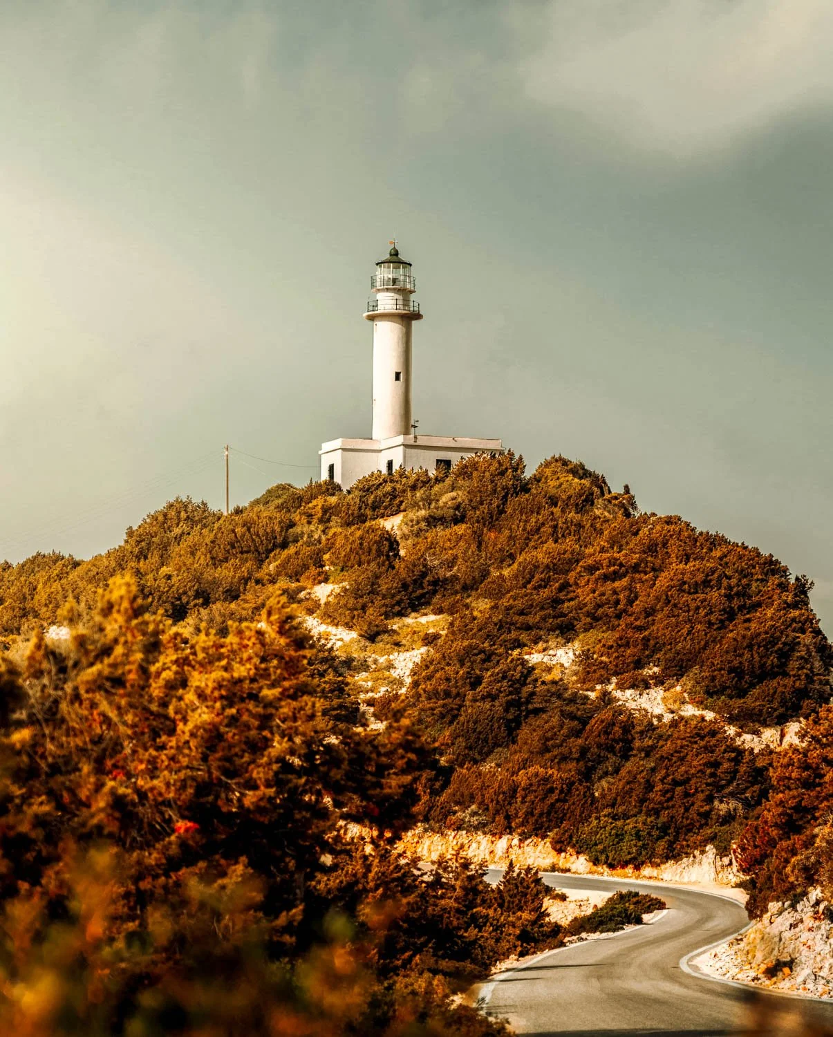 A white lighthouse atop a hill covered with brown and green bushes, next to a winding road under a cloudy sky.